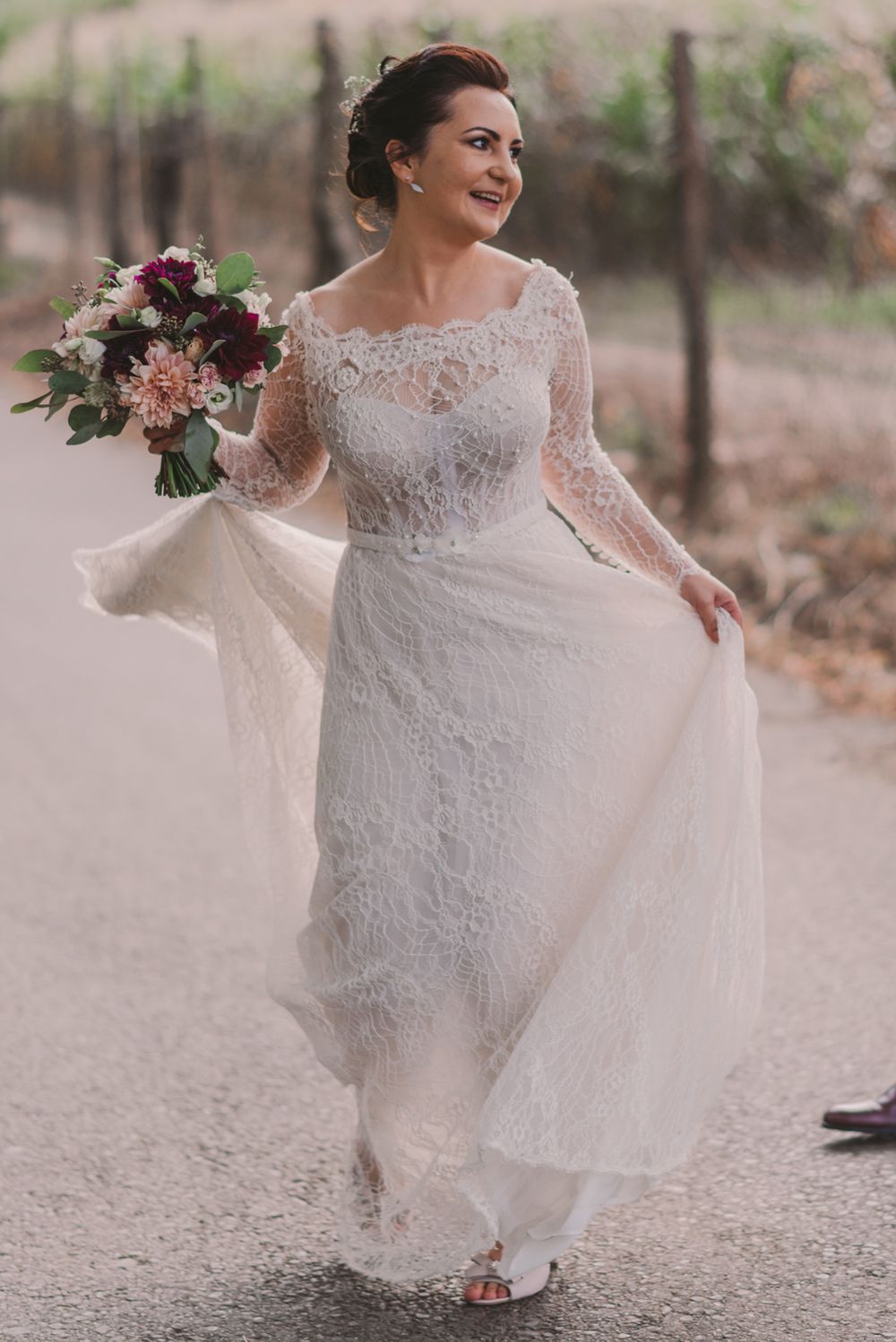 Bride in lace dress twirls playfully on a gravel path holding a pink and purple bouquet.