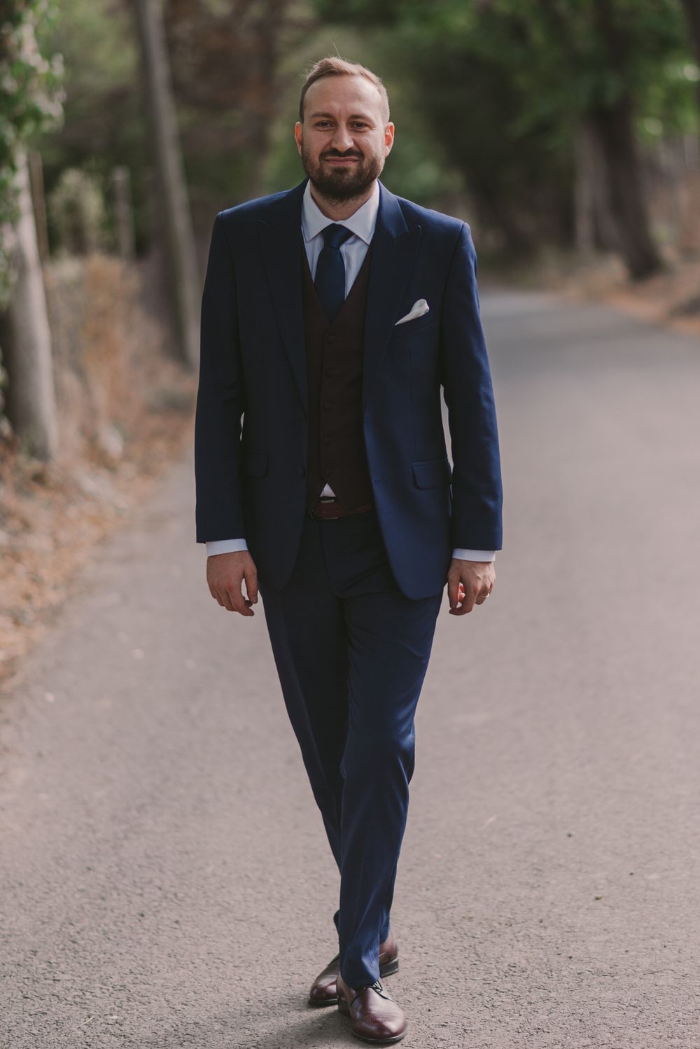 Groom in navy blue suit walks confidently down a tree-lined path during wedding photoshoot.