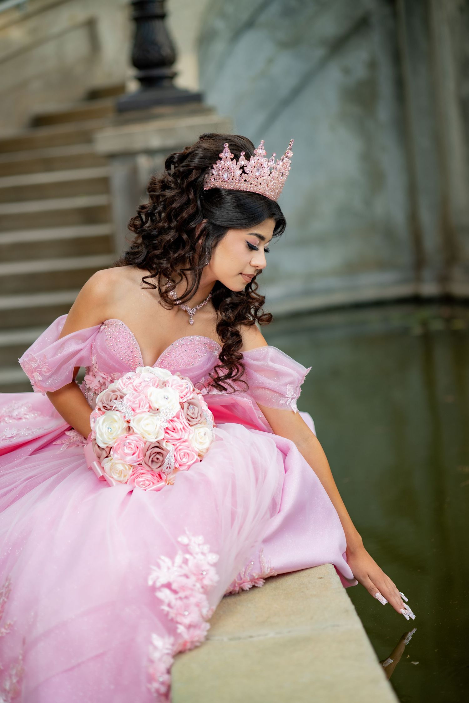 Young woman in pink quinceañera ball gown and crown holds bouquet while seated on stone ledge.
