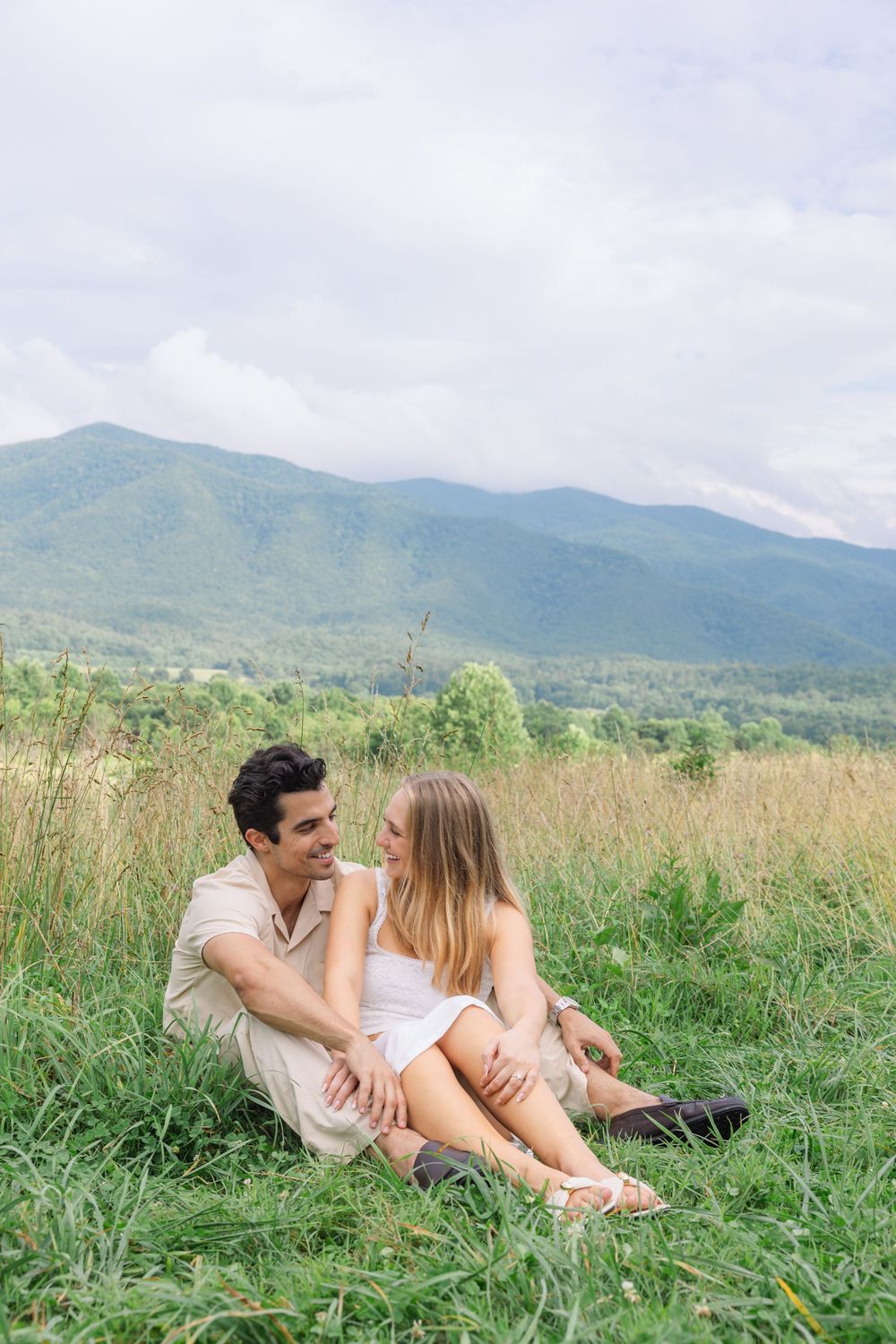 A couple sits together on a grassy hillside, smiling at each other with scenic green mountains in the background.