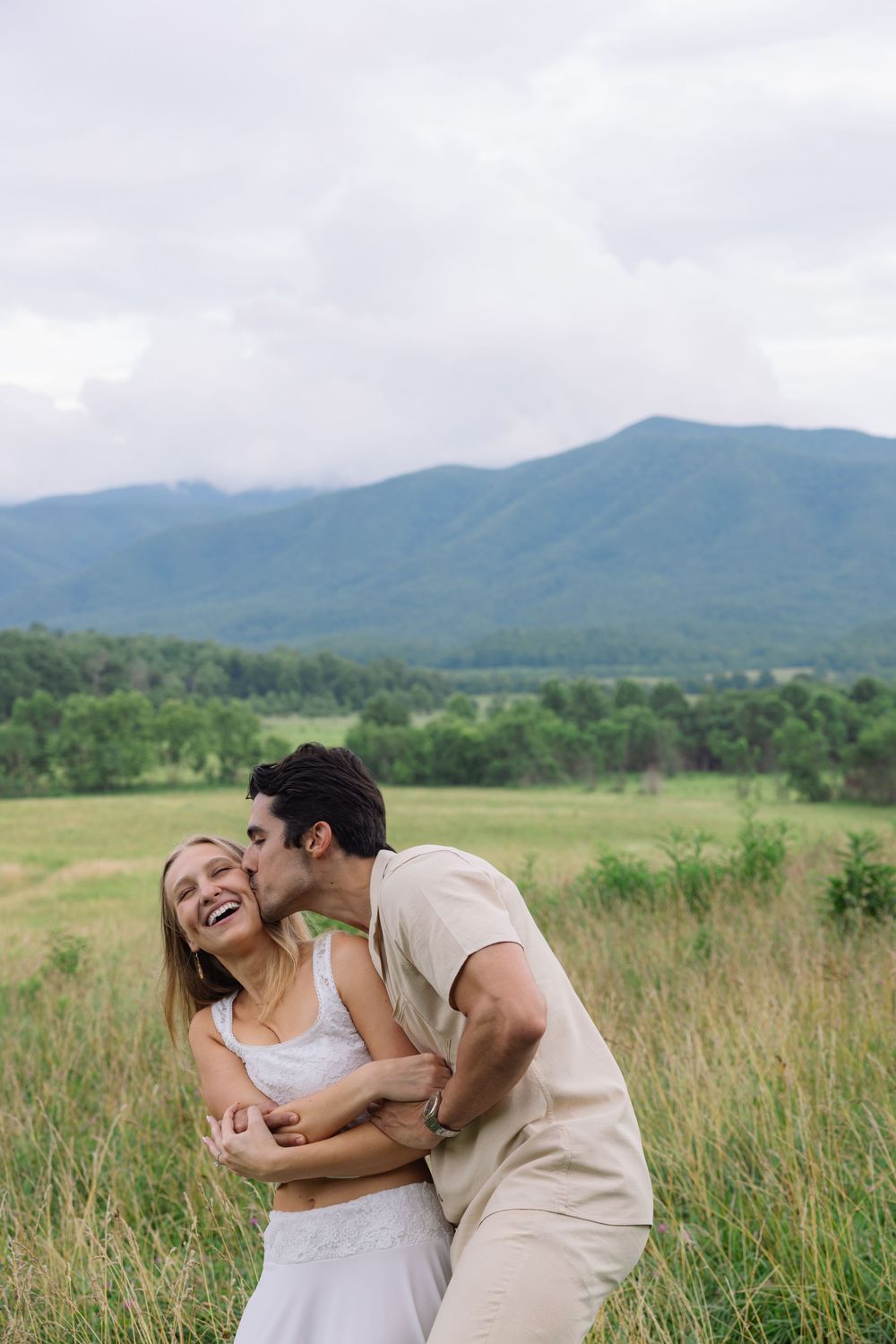 A couple embraces in a green meadow, man kissing woman's cheek, with mountains and cloudy sky in the background.
