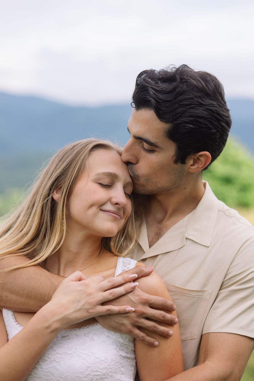 A man tenderly kisses a smiling woman's temple while embracing her from behind against a mountain backdrop.