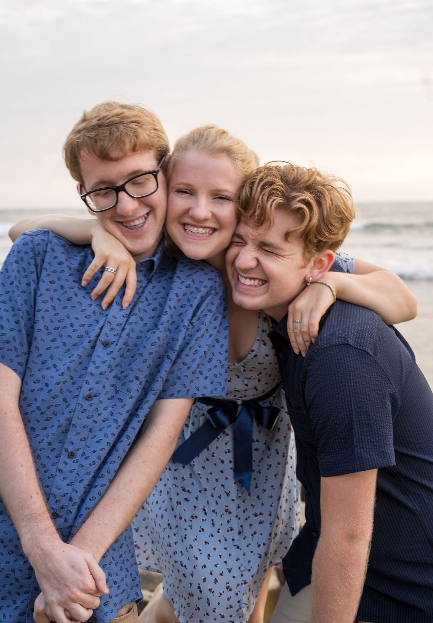 Three friends embrace and laugh together on a beach at sunset.