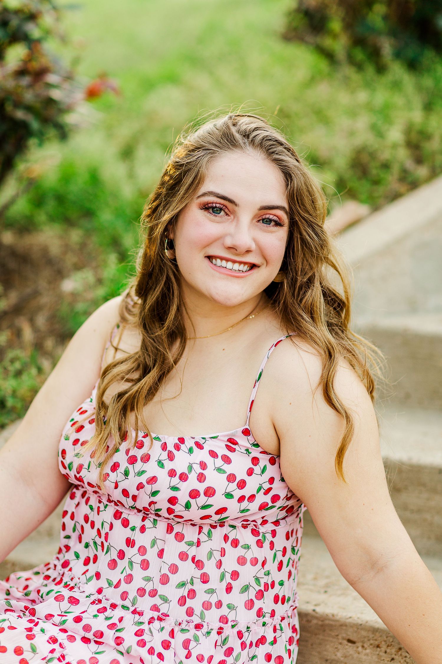 Graduate in cherry print dress poses outdoors with natural lighting and greenery.