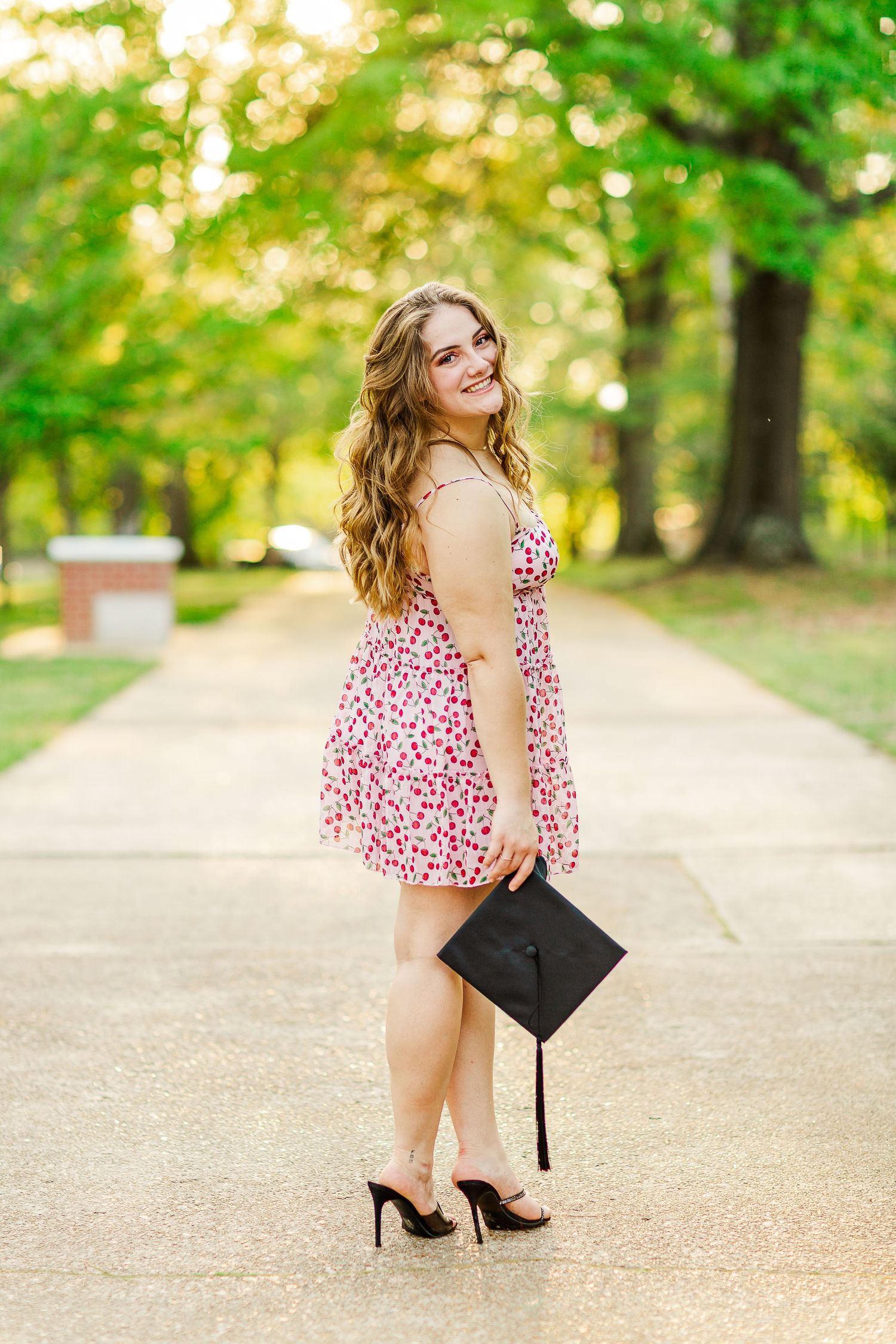Graduate in floral dress poses with graduation cap on campus walkway surrounded by green trees.