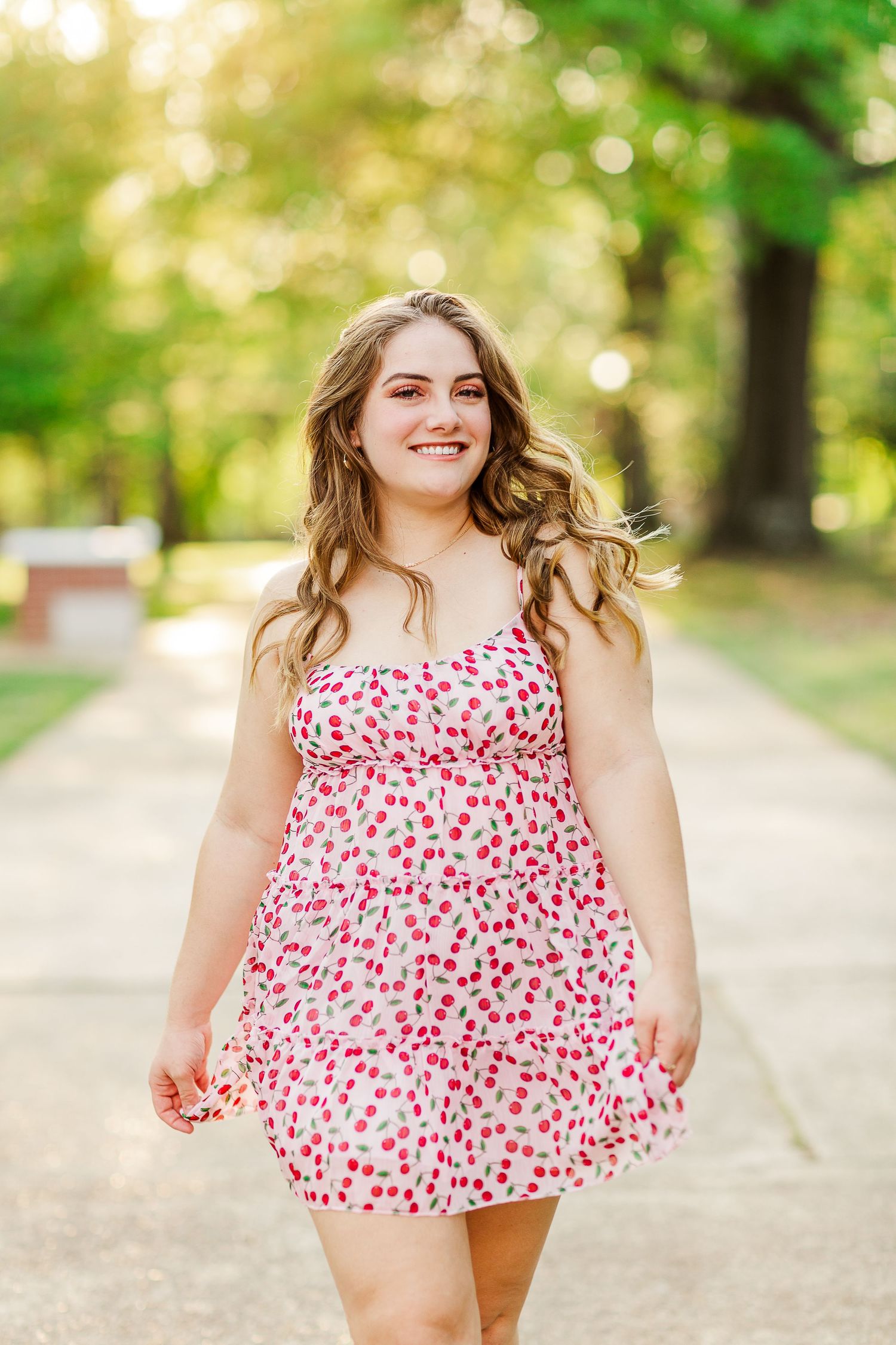 Graduate in floral print dress walks along path with natural bokeh background.