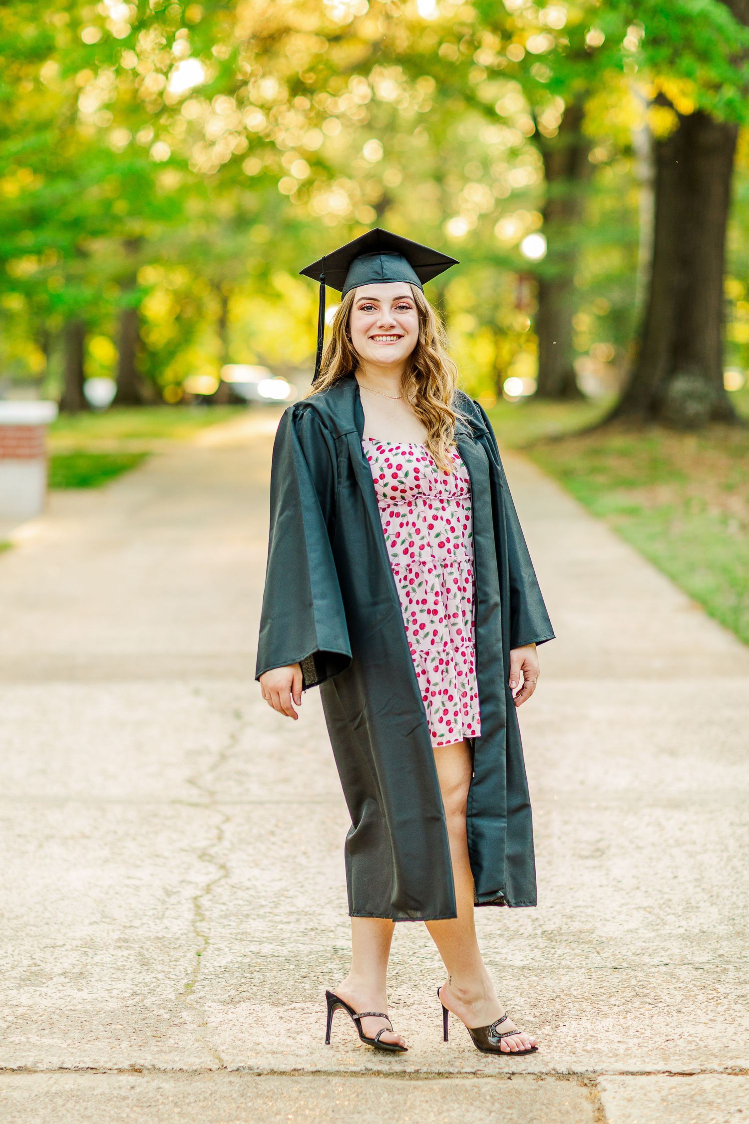 Full length shot of graduate in floral dress and black academic regalia on campus path.