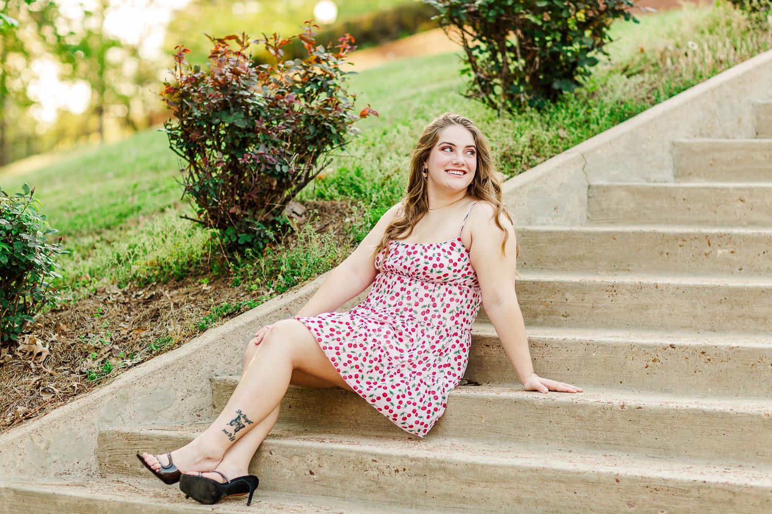 Graduate in floral dress sits on stone steps surrounded by landscaping on sunny day.