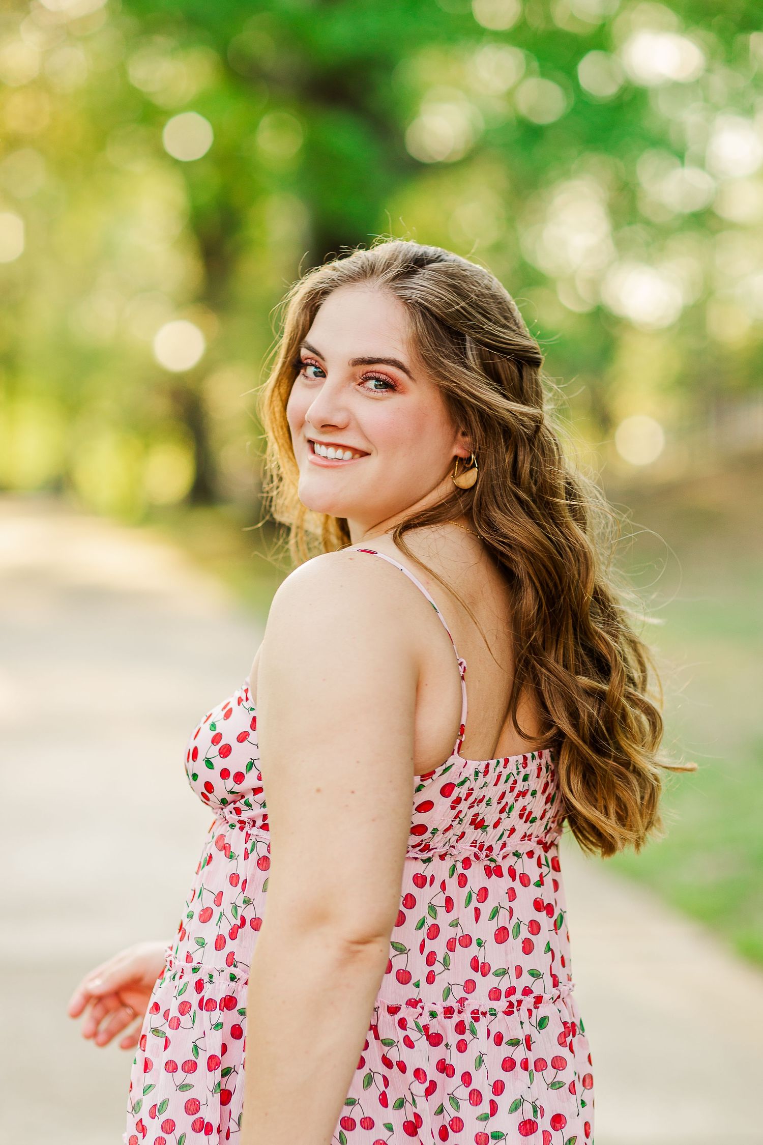 Graduate in cherry print dress poses outdoors with golden bokeh lighting effects.