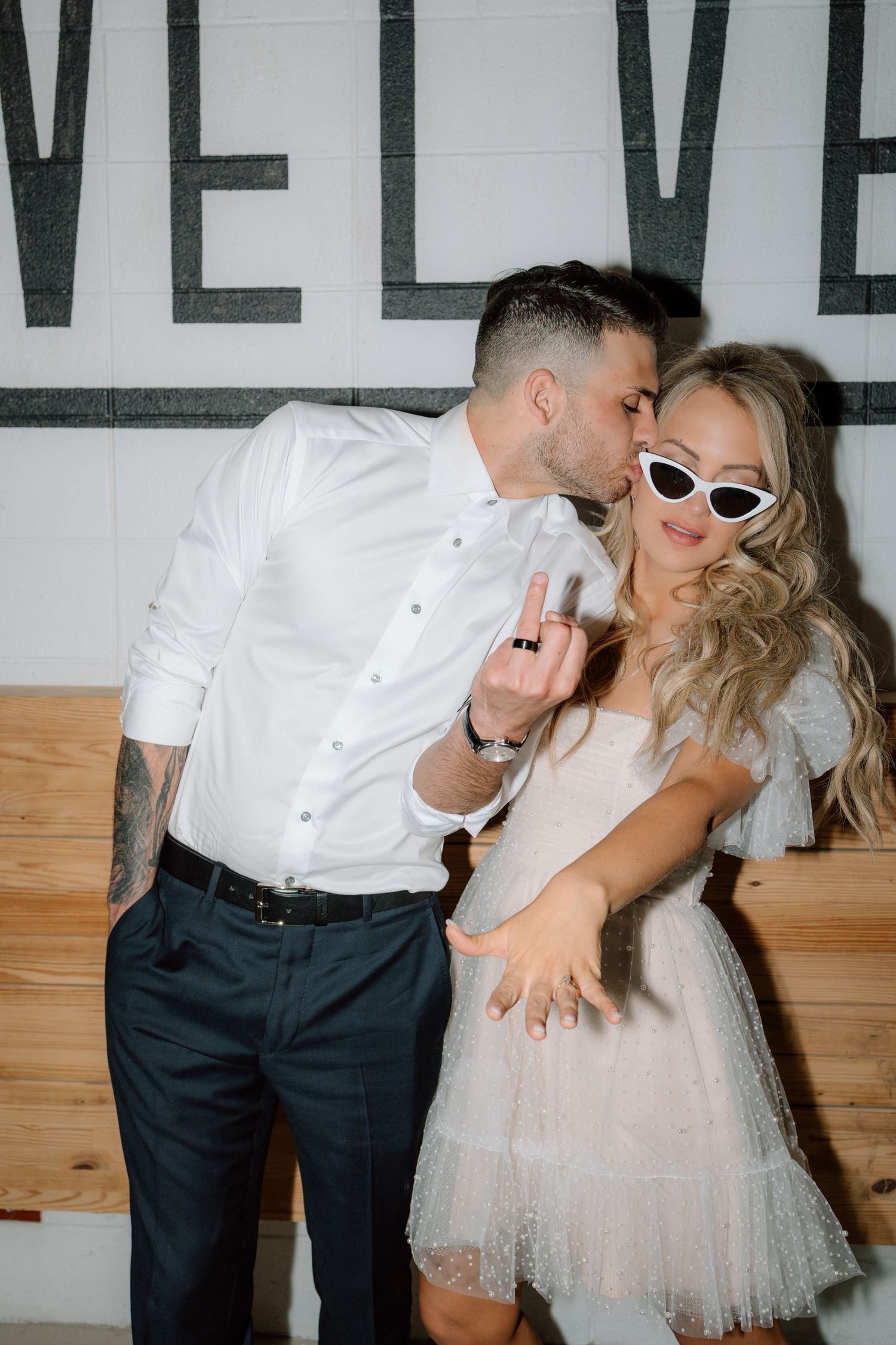 Playful couple in wedding attire pose against wall with 'LOVE' text, bride wearing retro sunglasses.