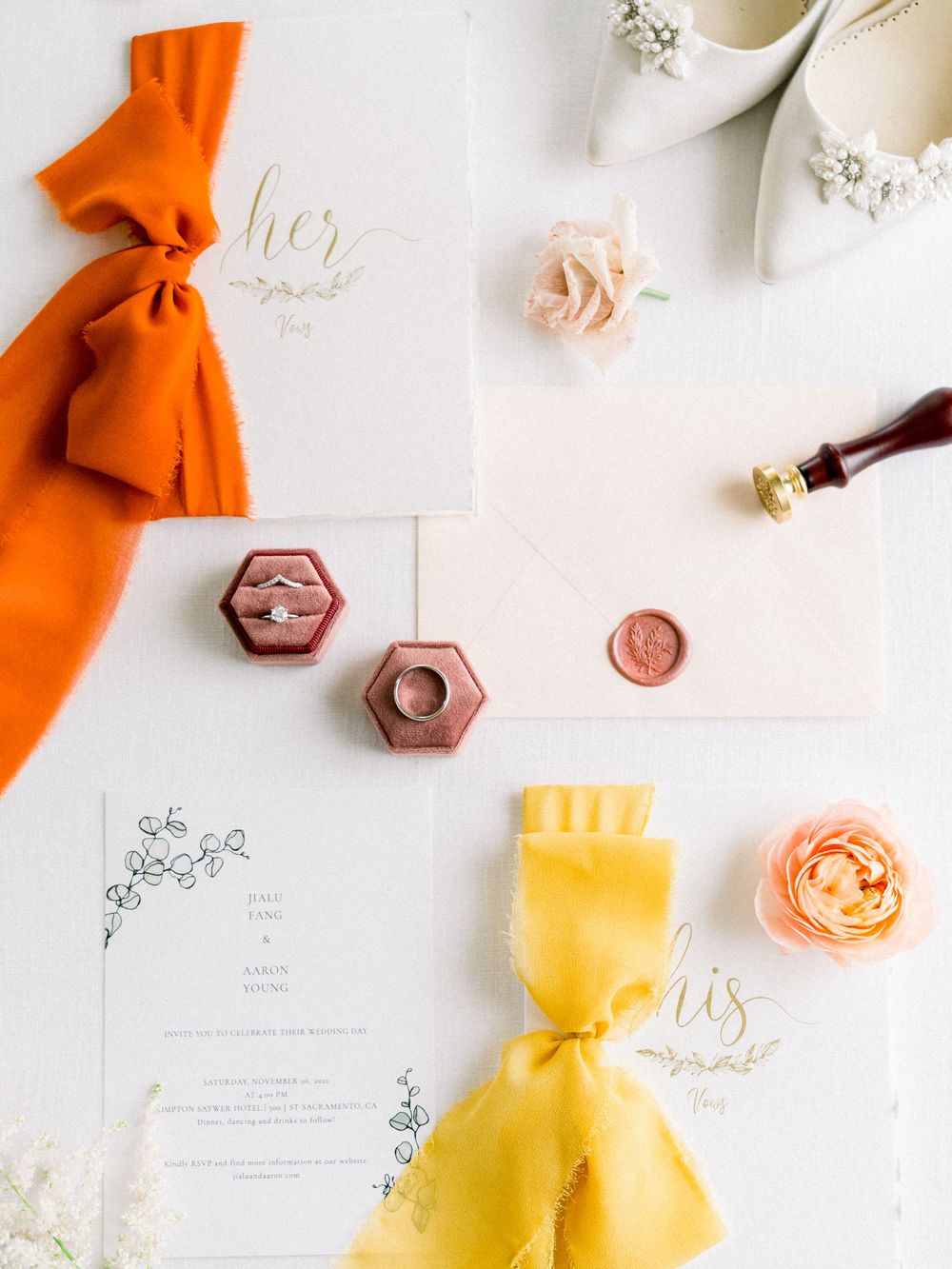 Wedding stationery flatlay with orange and yellow ribbons, wax seals, and hexagonal ring boxes.
