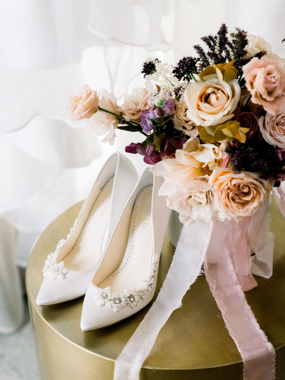 White embellished bridal shoes with romantic floral bouquet on gold table.