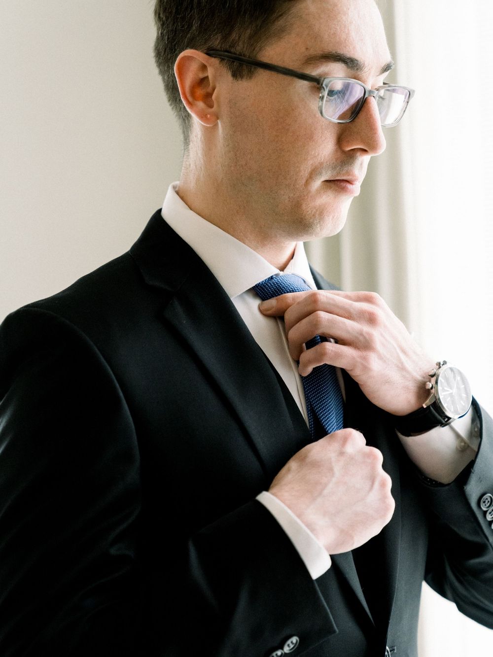 A person adjusting a blue tie while wearing a black suit and glasses.