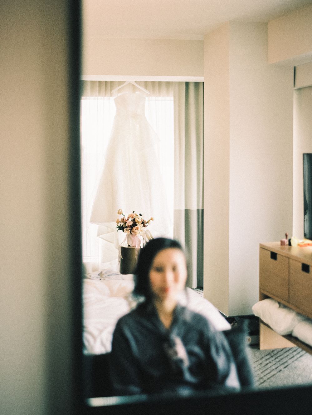 A white wedding dress hangs by a window with a floral arrangement in a hotel room.
