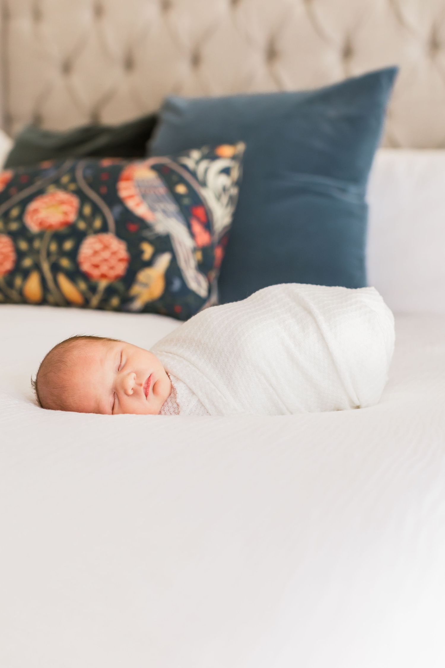 A sleeping newborn baby wrapped in white swaddle on a bed with decorative pillows.
