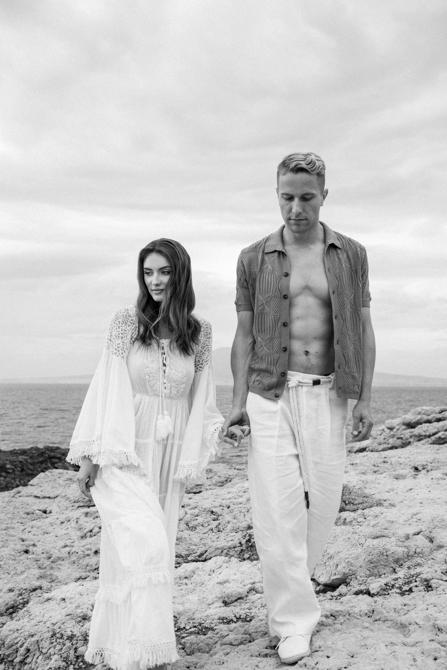 Black and white photo of couple in casual white outfits standing on rocky coastal terrain.