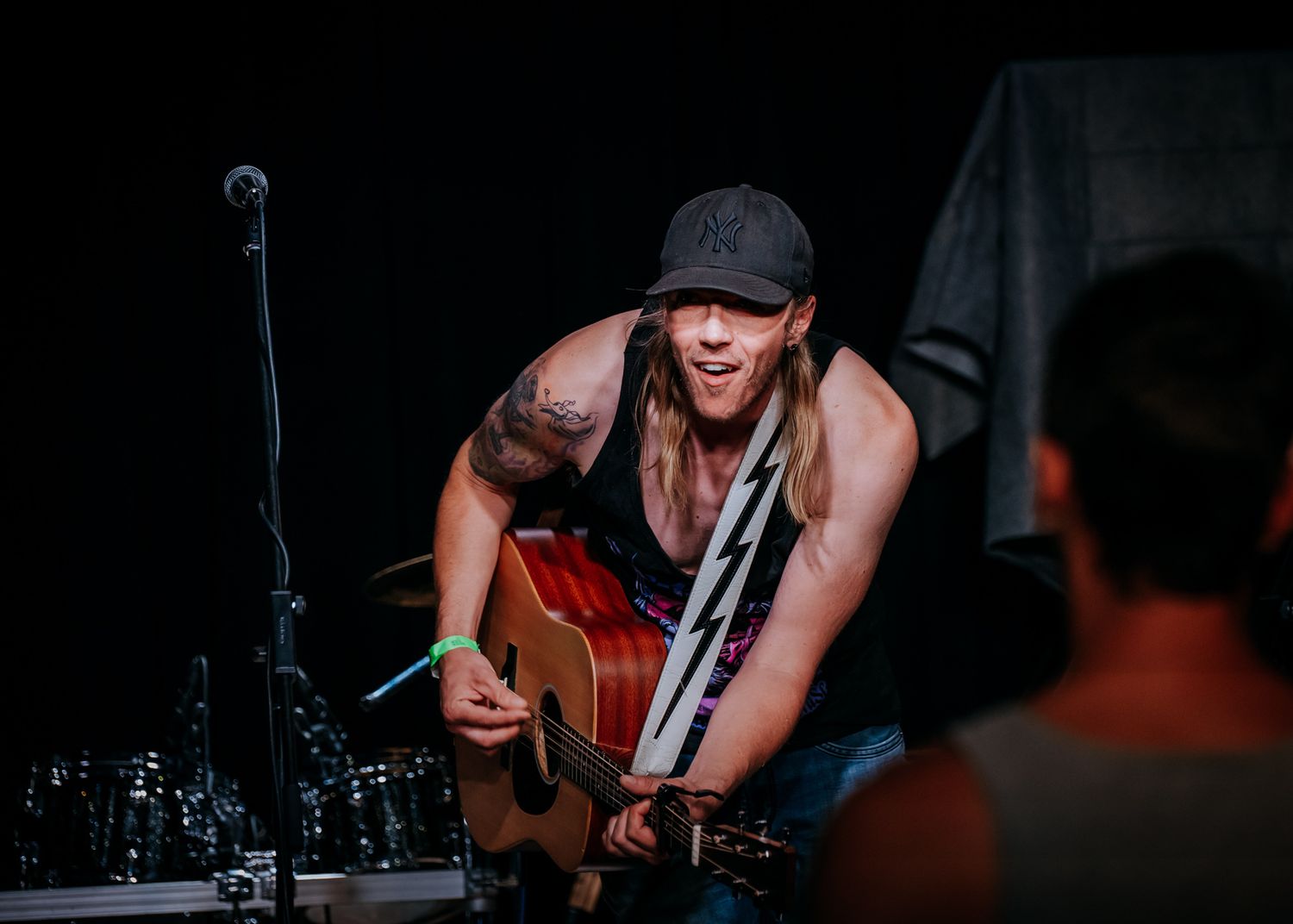 Musician in sleeveless shirt and cap plays acoustic guitar on dark stage with microphone setup.