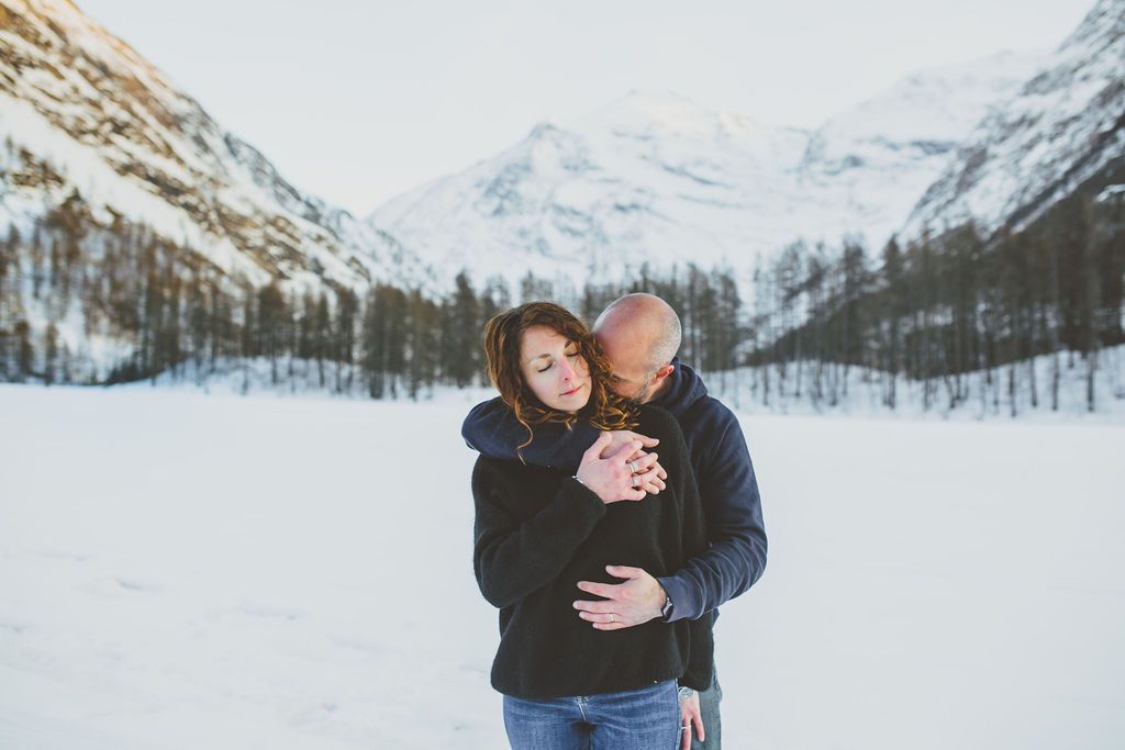 A couple embraces in a snowy winter mountain landscape with snow-covered trees in the background.