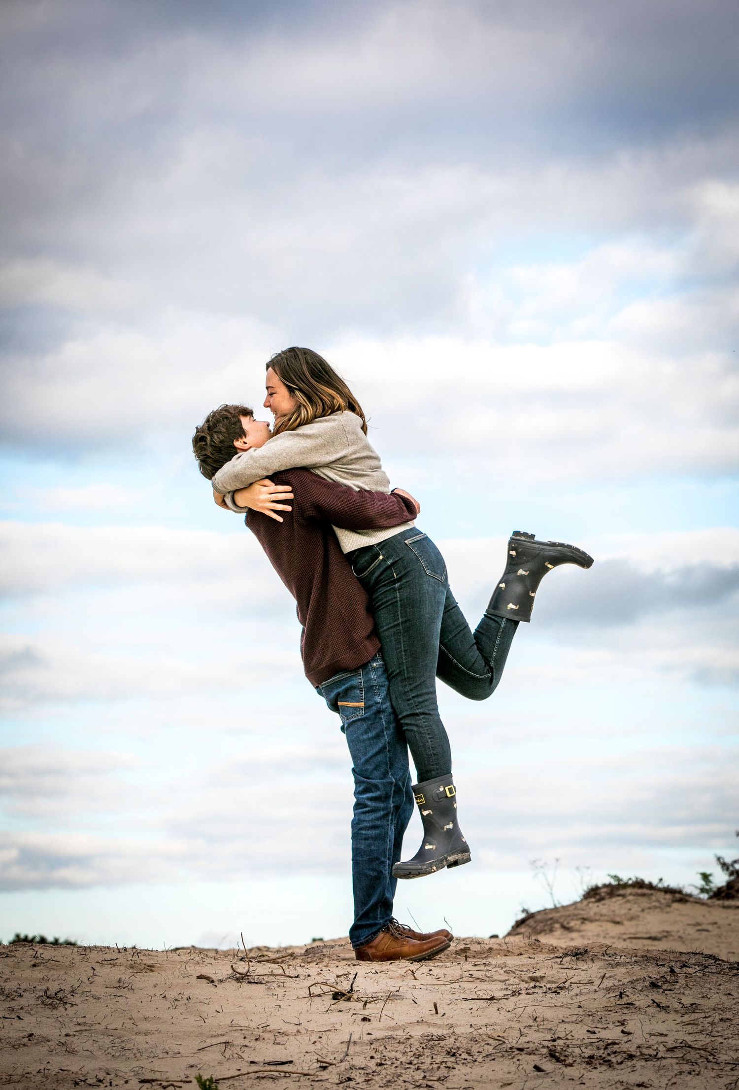 Playful couple embracing and lifting each other on a sandy hill against cloudy sky.