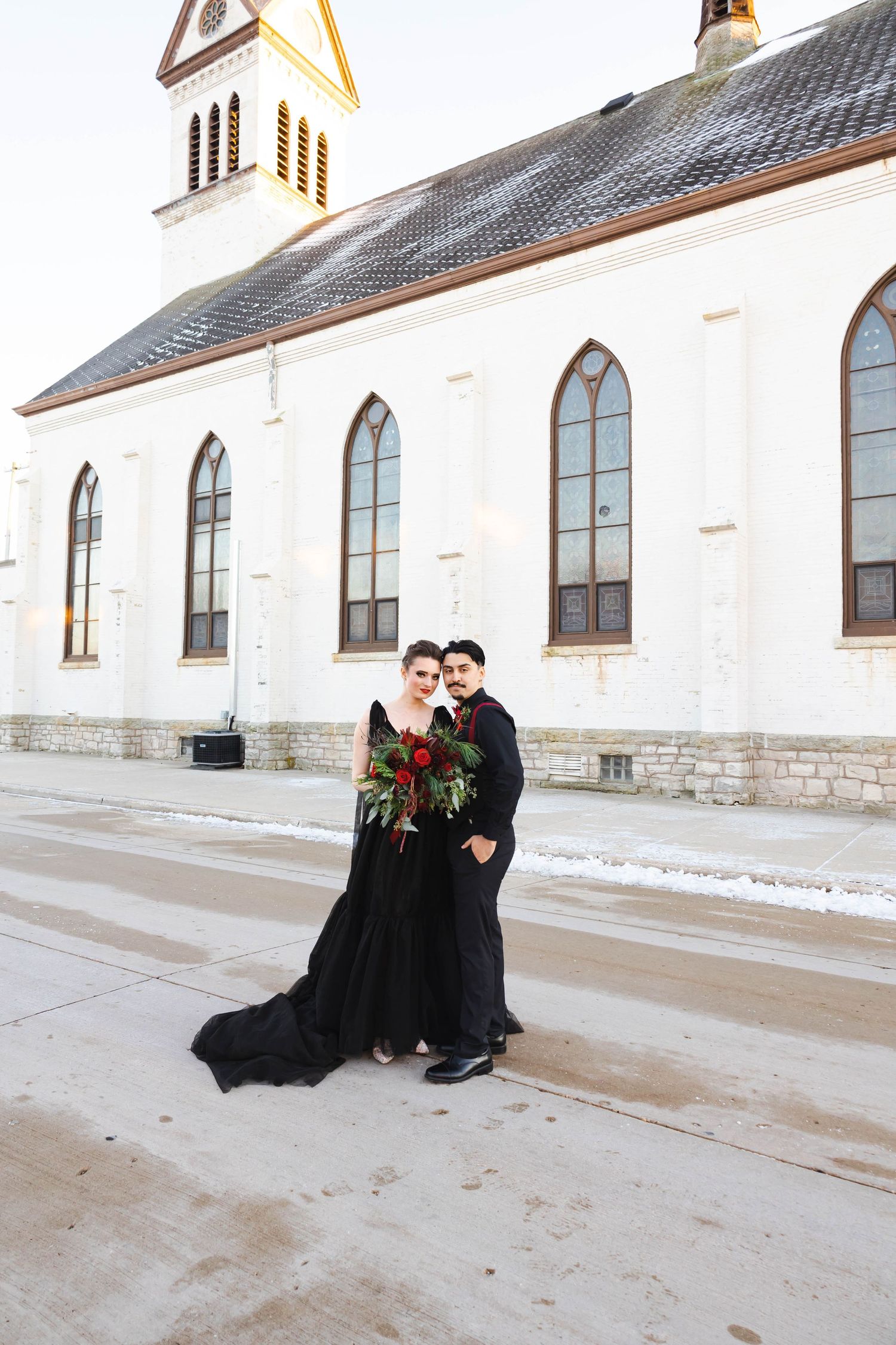 Couple in formal attire posing outside white church with dramatic architecture.