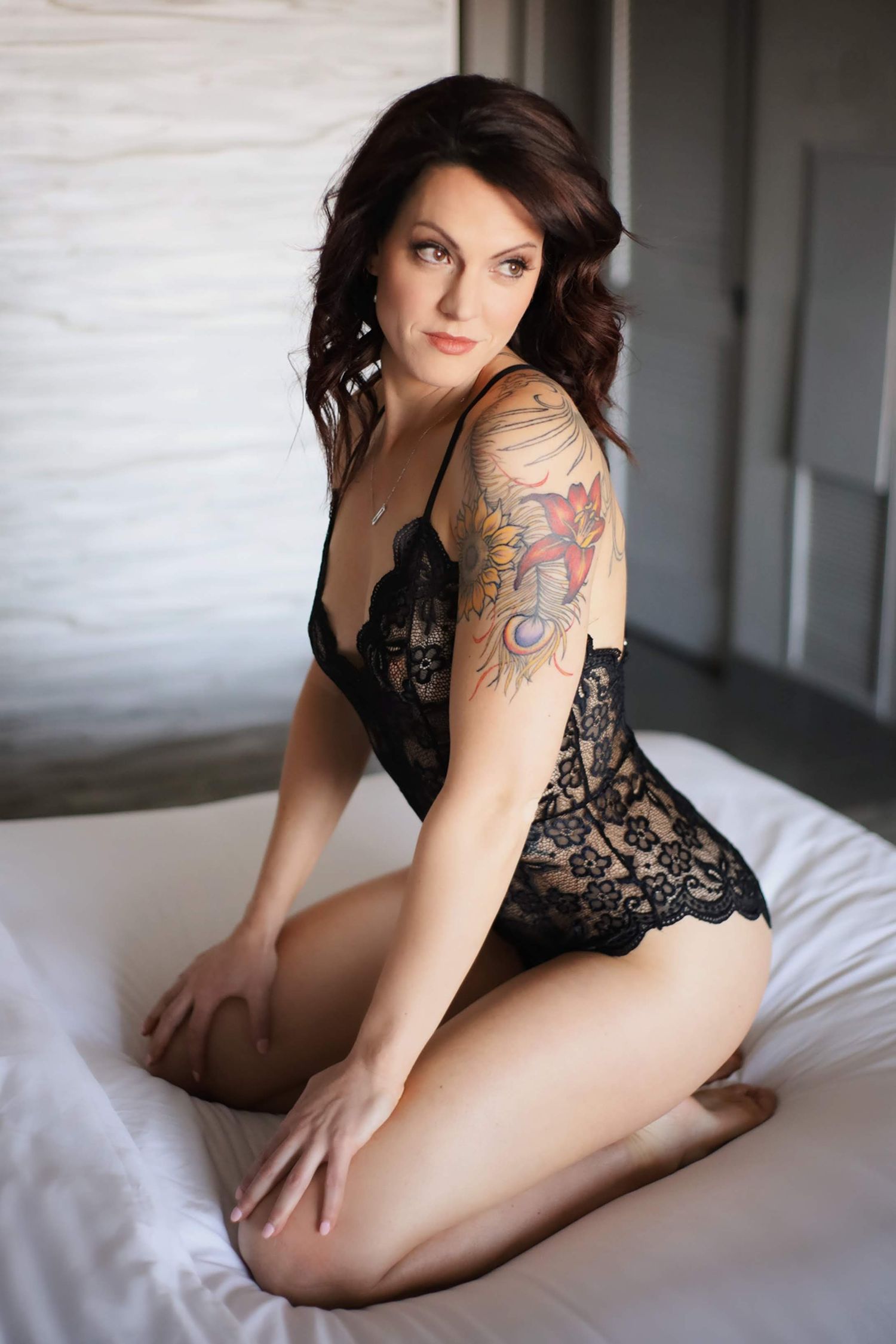 Woman in black lace lingerie poses on white bedding with visible arm tattoo.