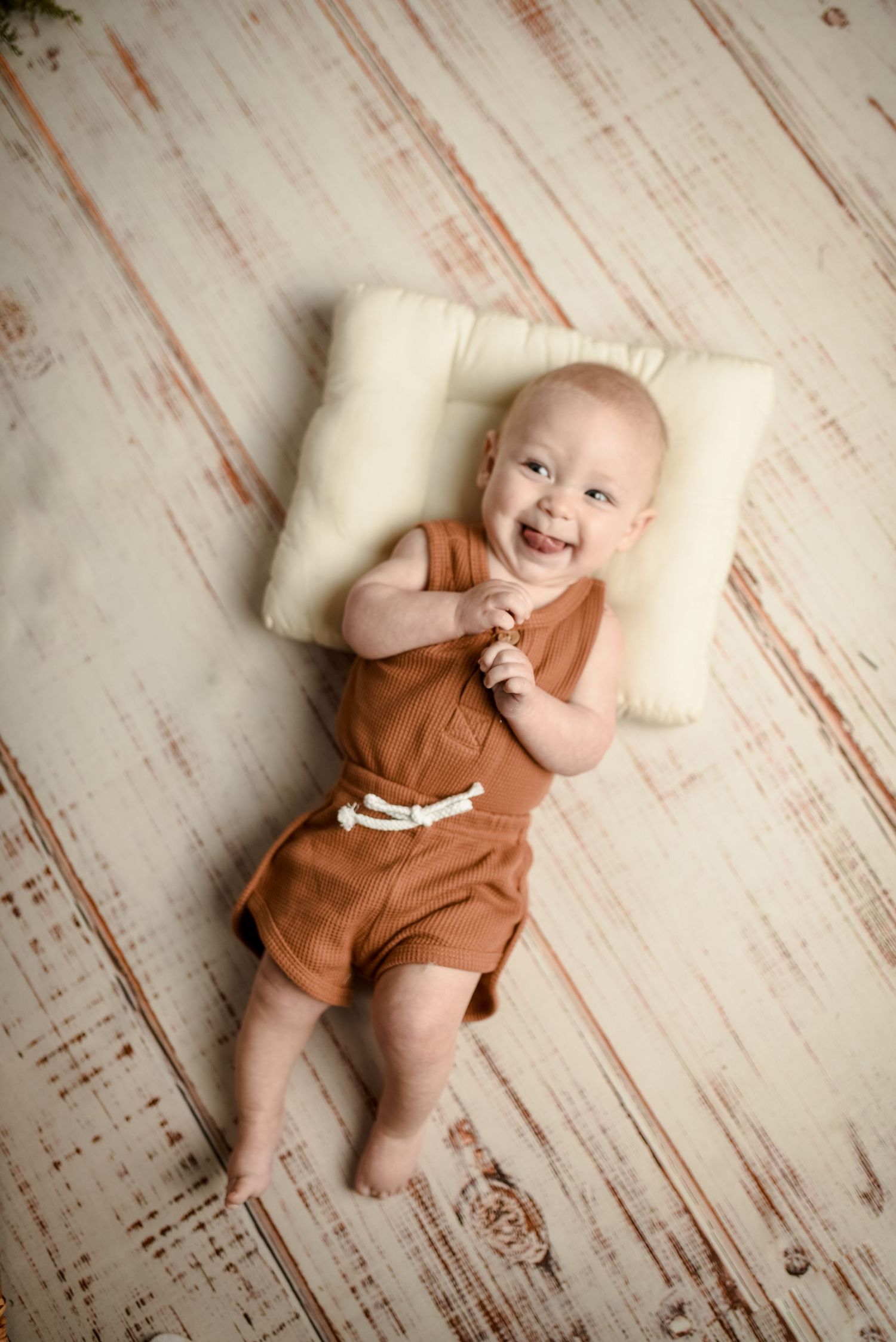 Baby in brown romper lying on white pillow against distressed wooden background.