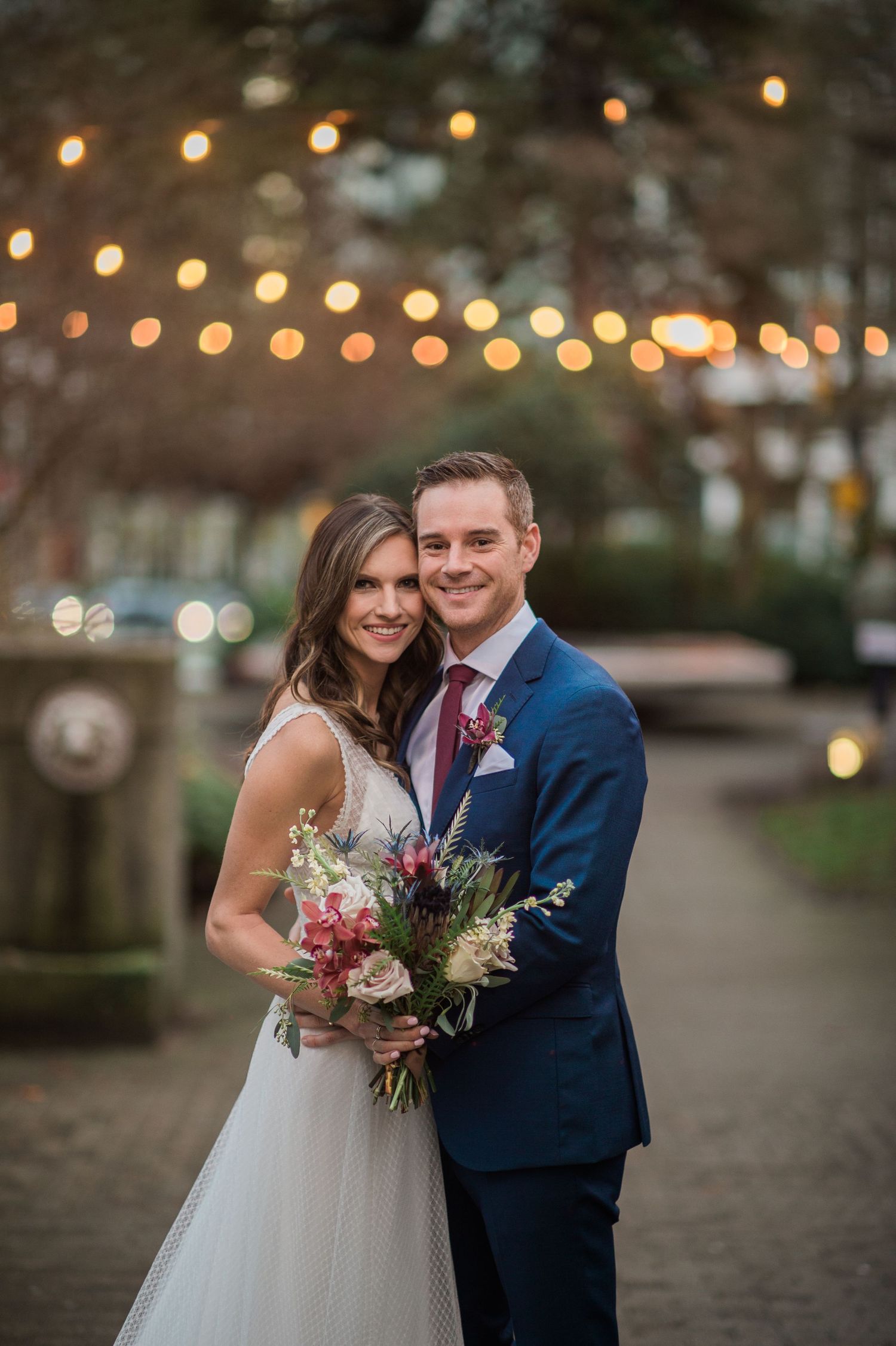 A romantic wedding photo under string lights at dusk with beautiful floral bouquet.