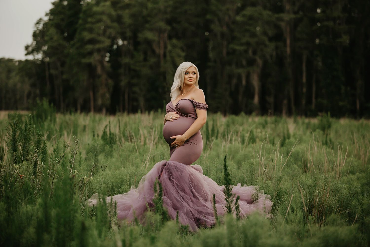Maternity portrait in a purple flowing gown amid tall grass and forest backdrop.