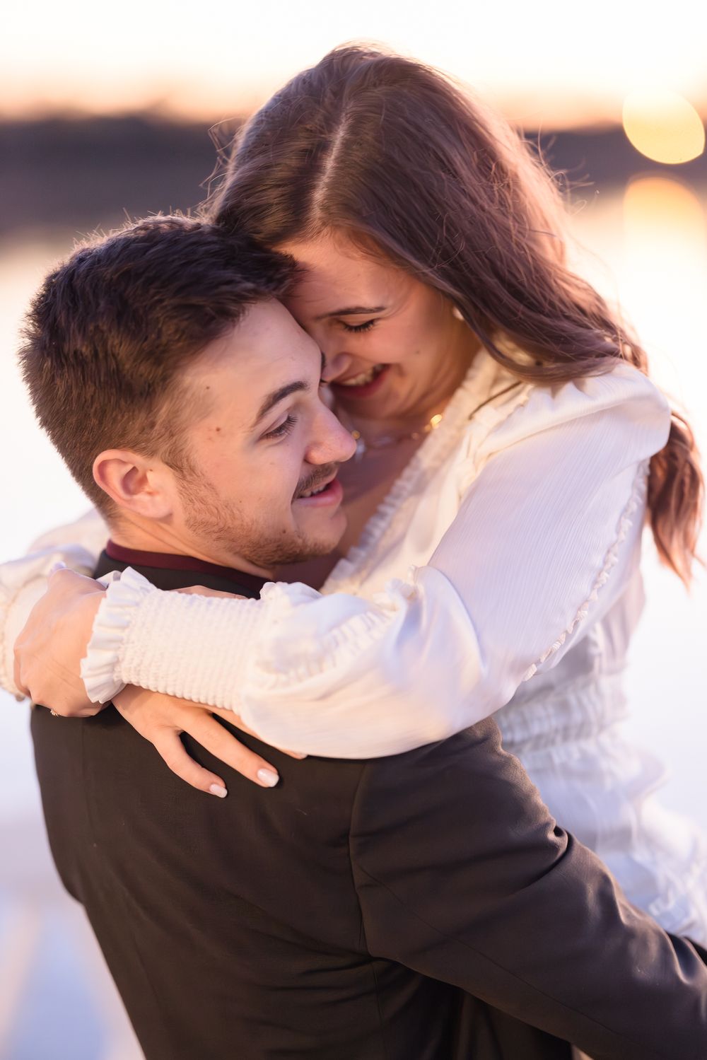 Romantic sunset photoshoot with couple embracing in casual attire by waterfront.