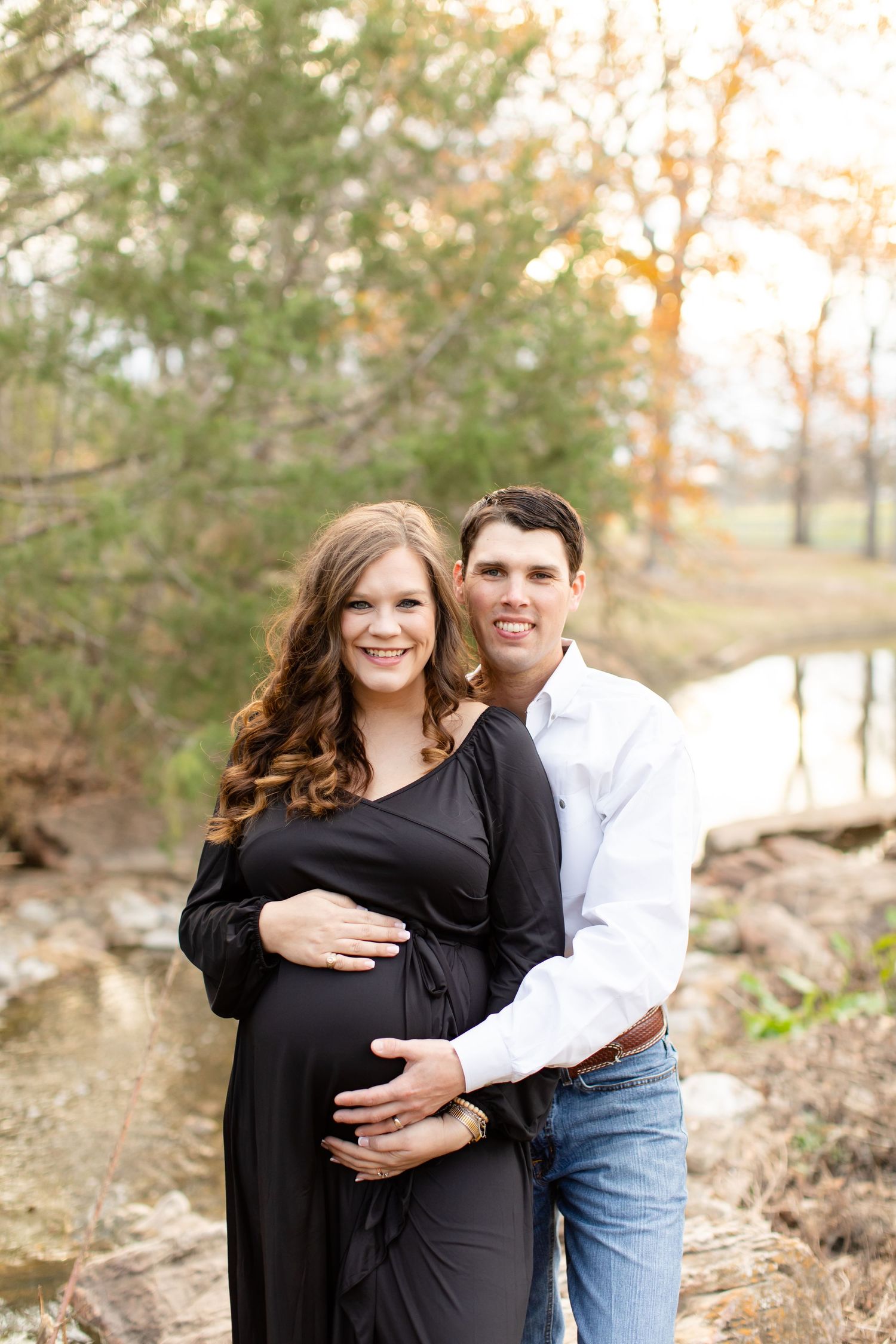 Maternity portrait by creek with couple in black dress and white shirt.