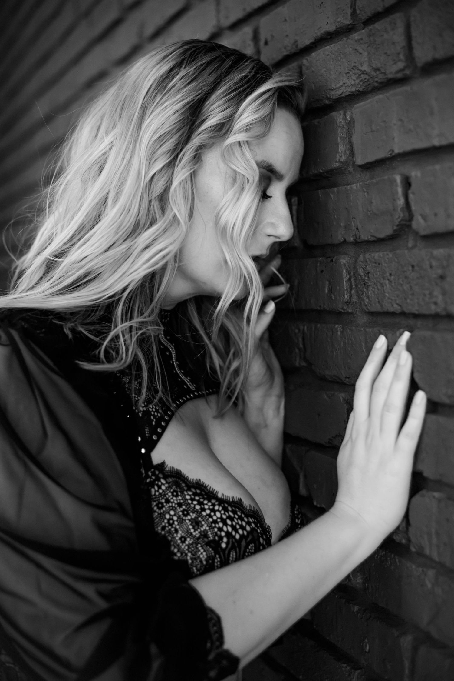 Black and white portrait of woman with wavy hair leaning against brick wall.