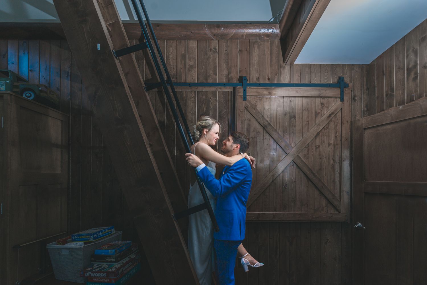 A couple embraces romantically by a rustic wooden barn door in dramatic lighting.
