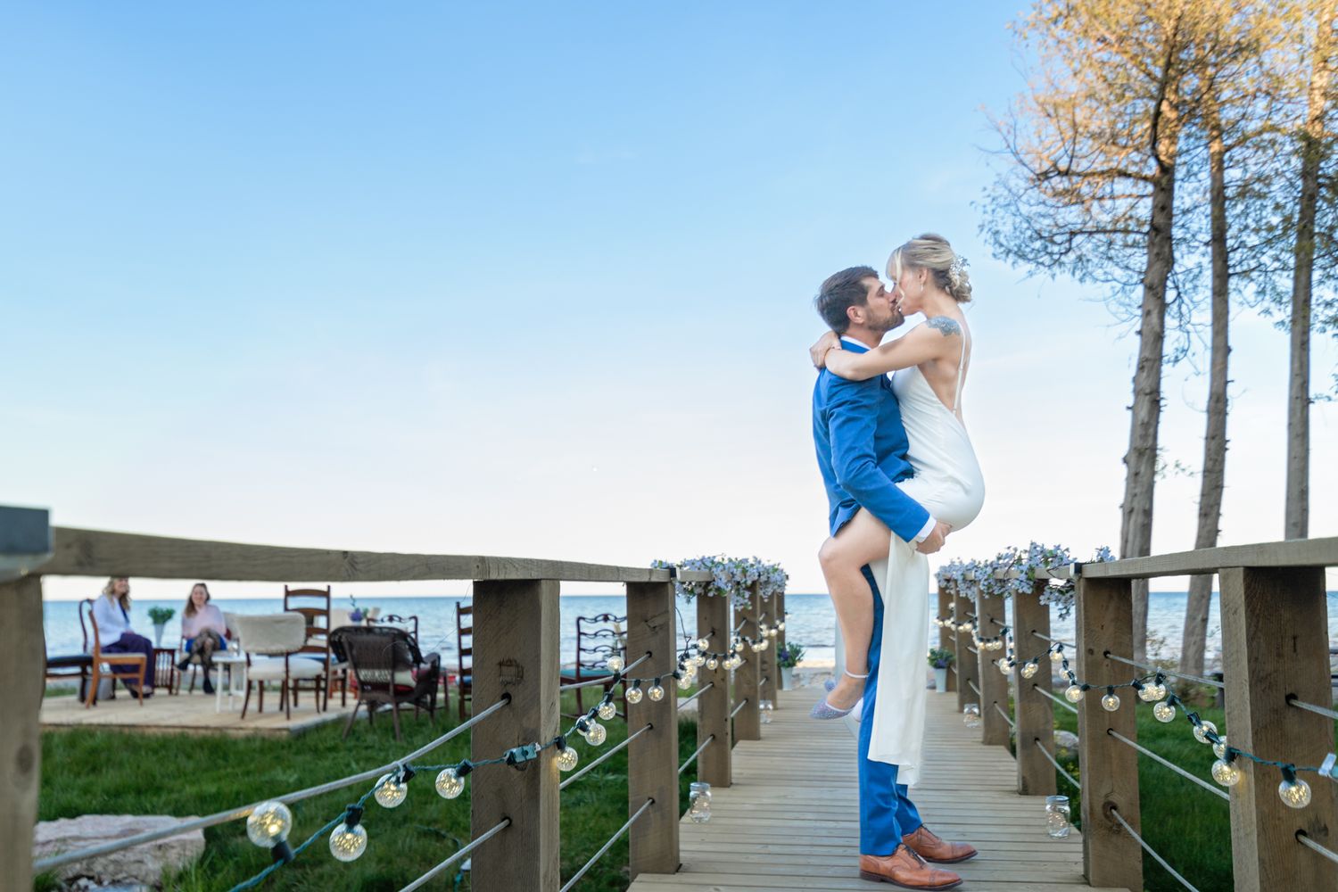 A romantic moment captured on a decorated wooden deck at sunset with white lights and tall evergreen trees.