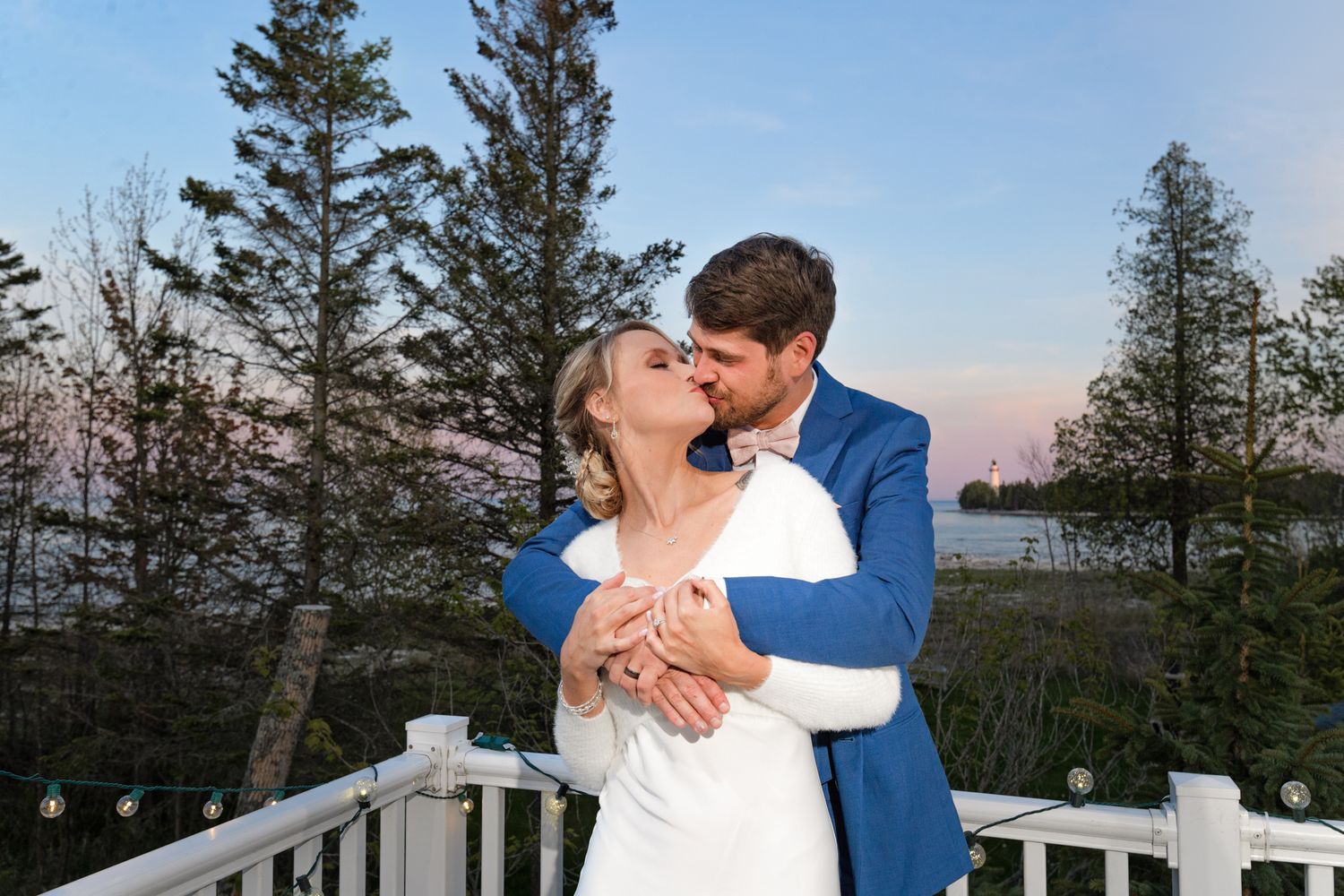 A romantic embrace on a white railed deck with evergreen trees and water in the background at dusk.