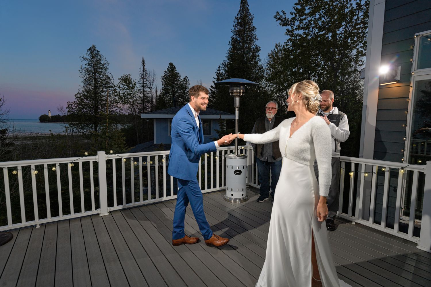People dancing on an outdoor deck at dusk with string lights and pine trees in the background.
