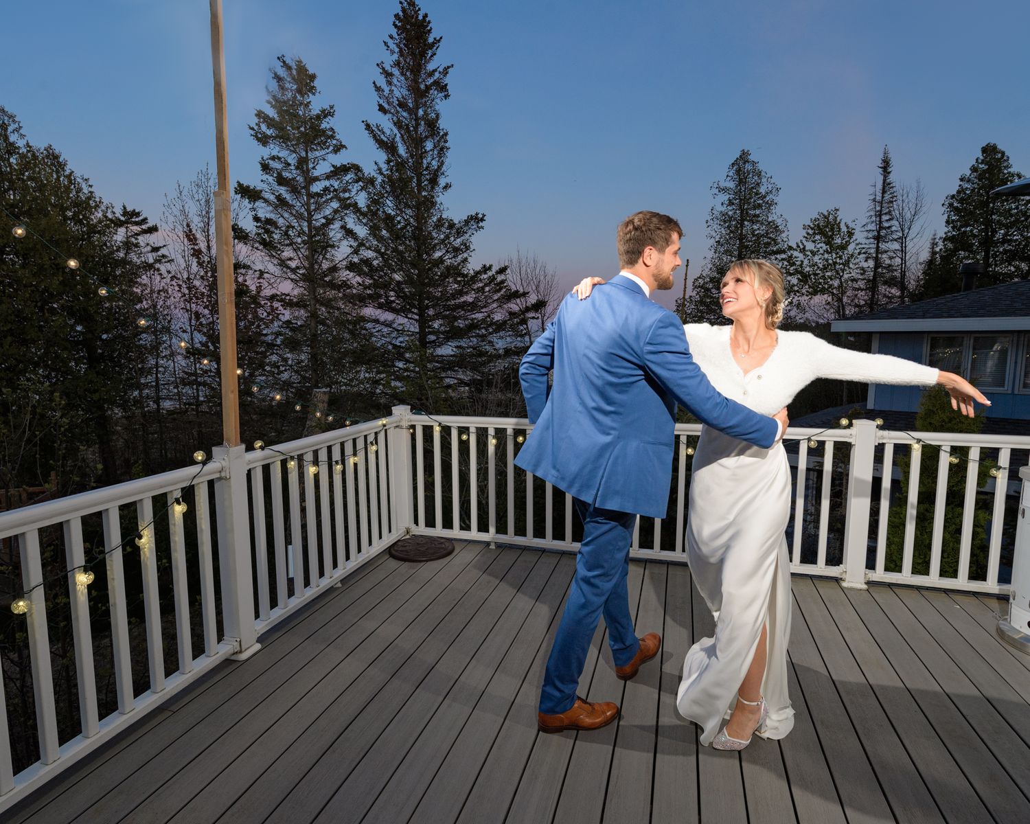A dancing couple in formal attire performs elegant moves on a wooden deck surrounded by evergreen trees.