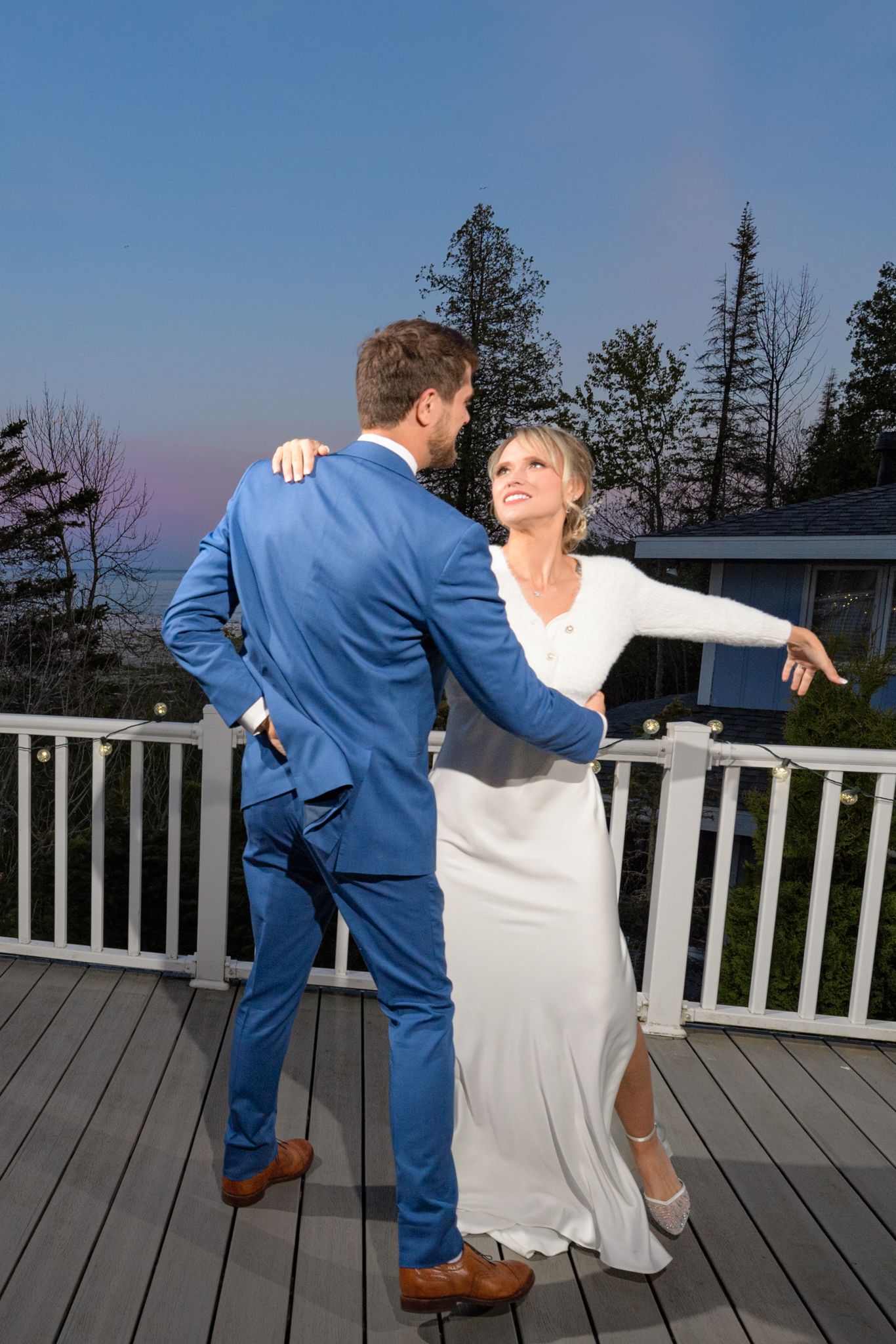 A couple dances intimately on an outdoor deck at sunset with pine trees and white railings in the background.