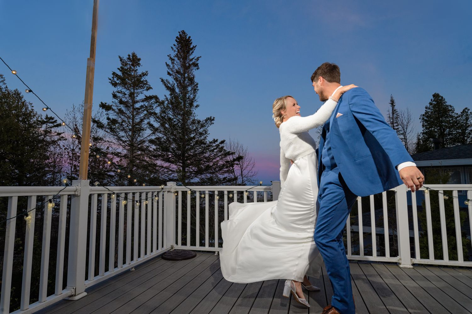 A graceful dancing pose on a wooden deck with evergreen trees silhouetted against the evening sky.