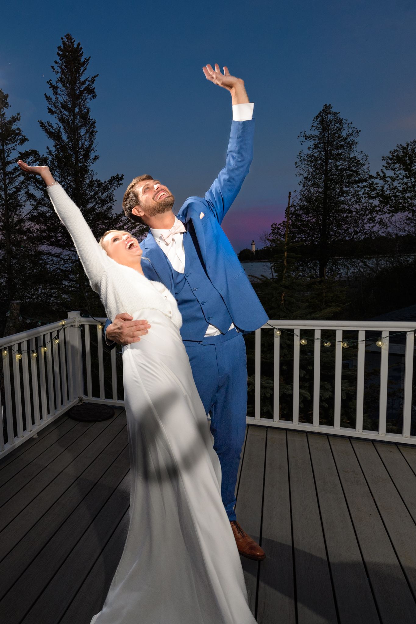 Wedding couple striking dramatic pose with raised arms on outdoor deck at dusk with evergreen trees in background.