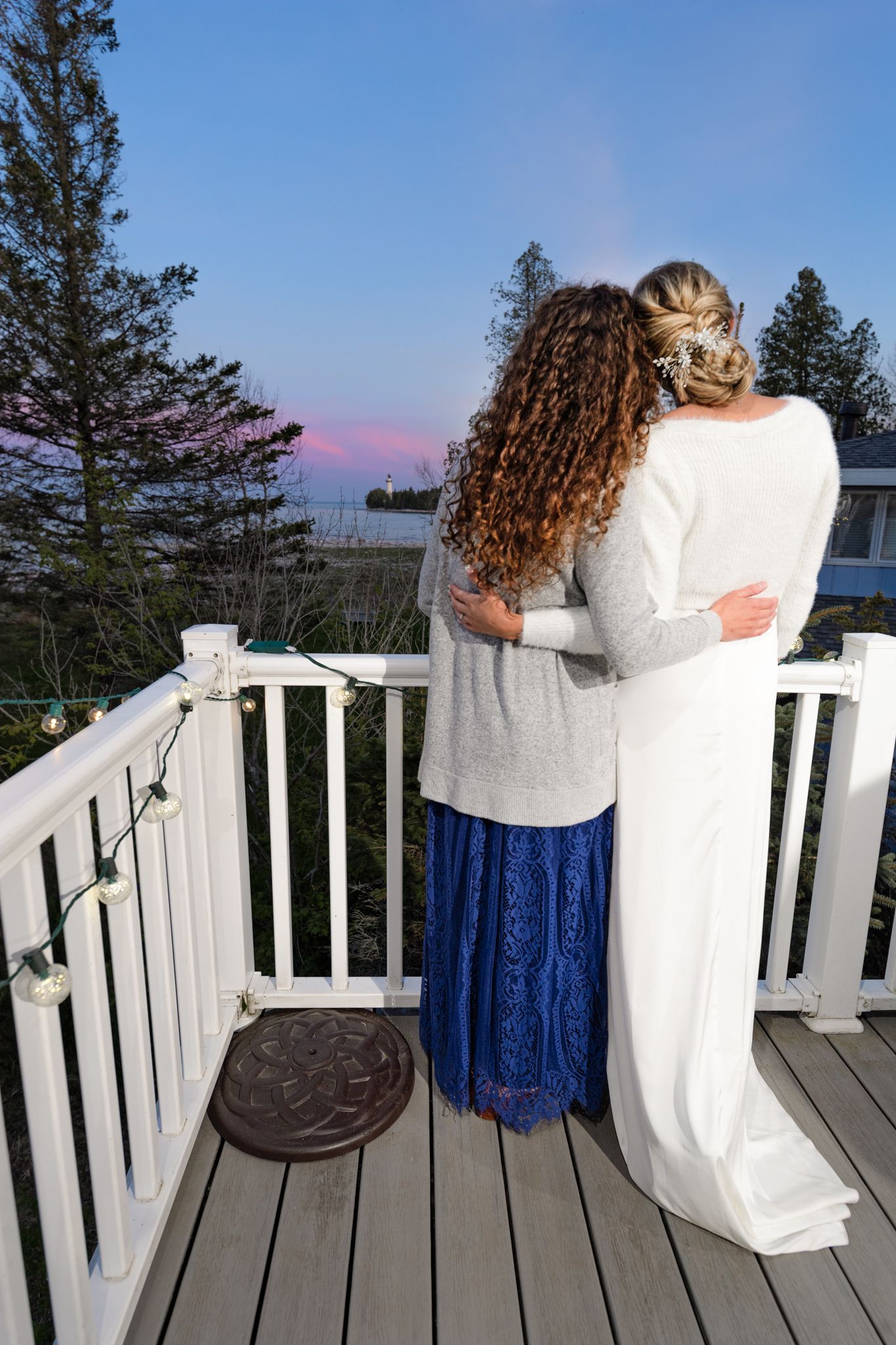 Two people embrace while watching sunset from a deck overlooking water and pine trees.