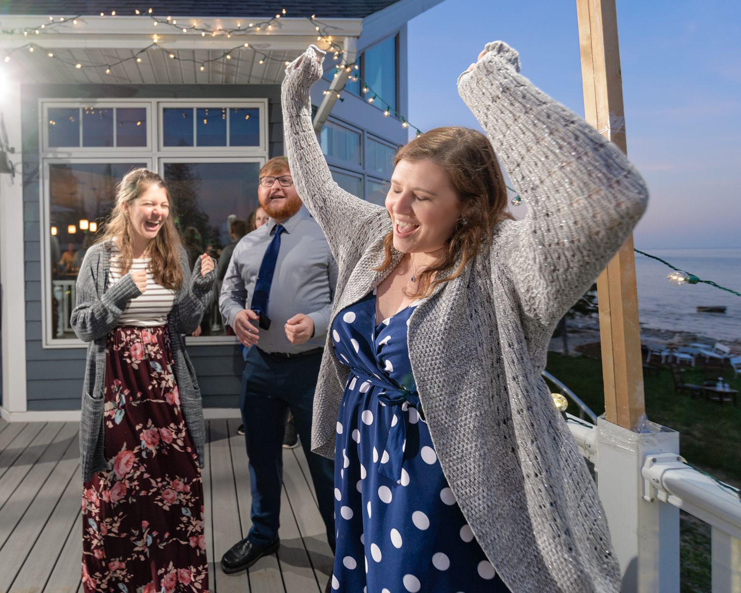 Celebration on outdoor deck with string lights as people dance and socialize.