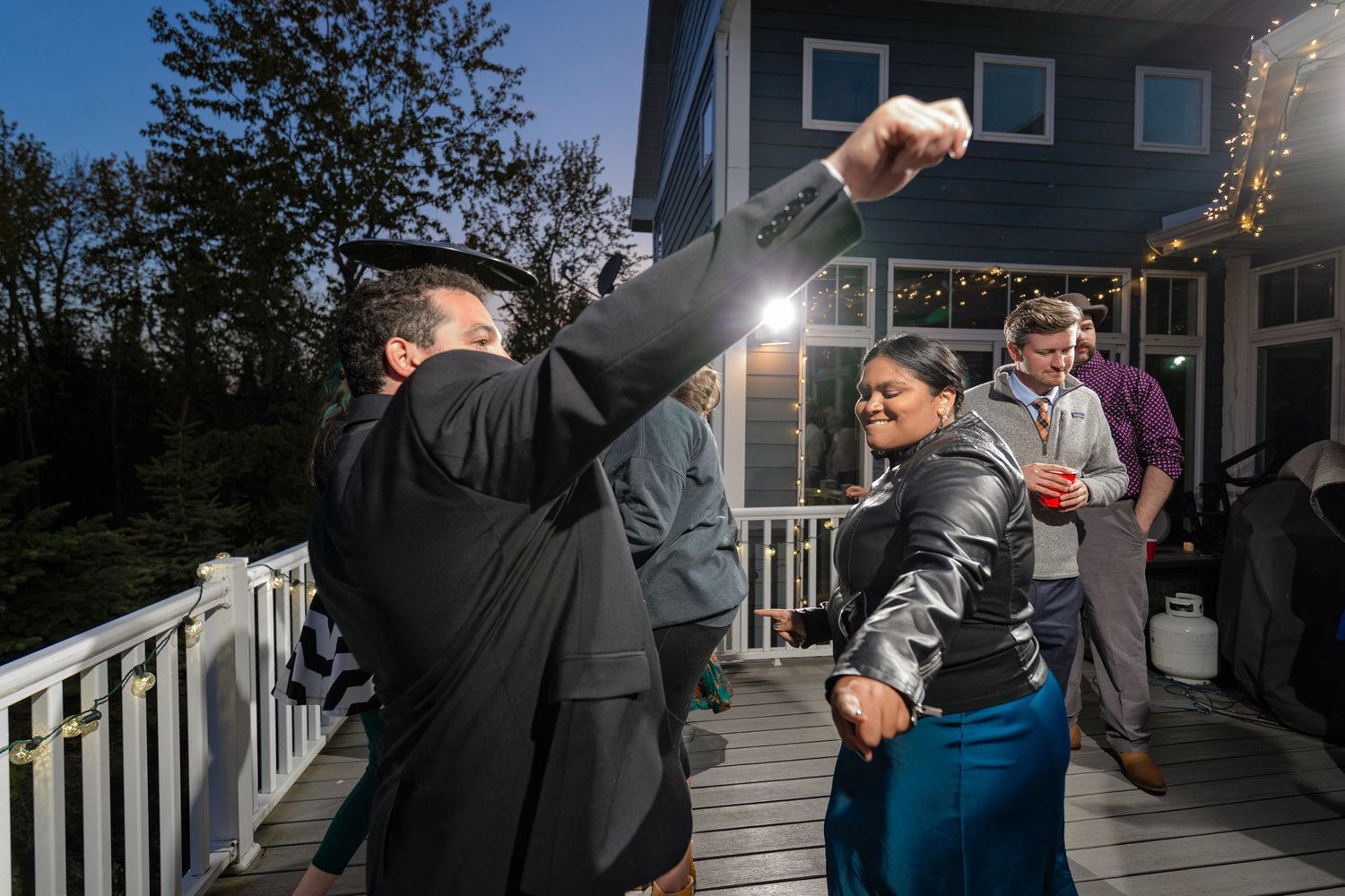 Wedding party dancing energetically on deck with string lights and trees in background.