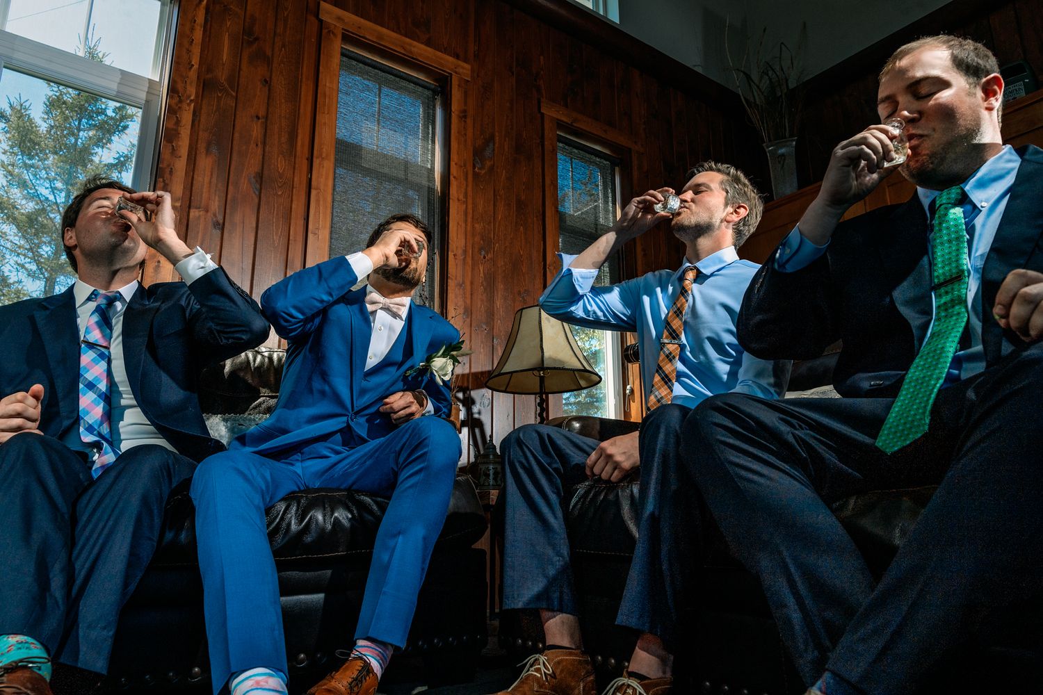 Groomsmen in suits share drinks and laughs on a porch during a wedding celebration.