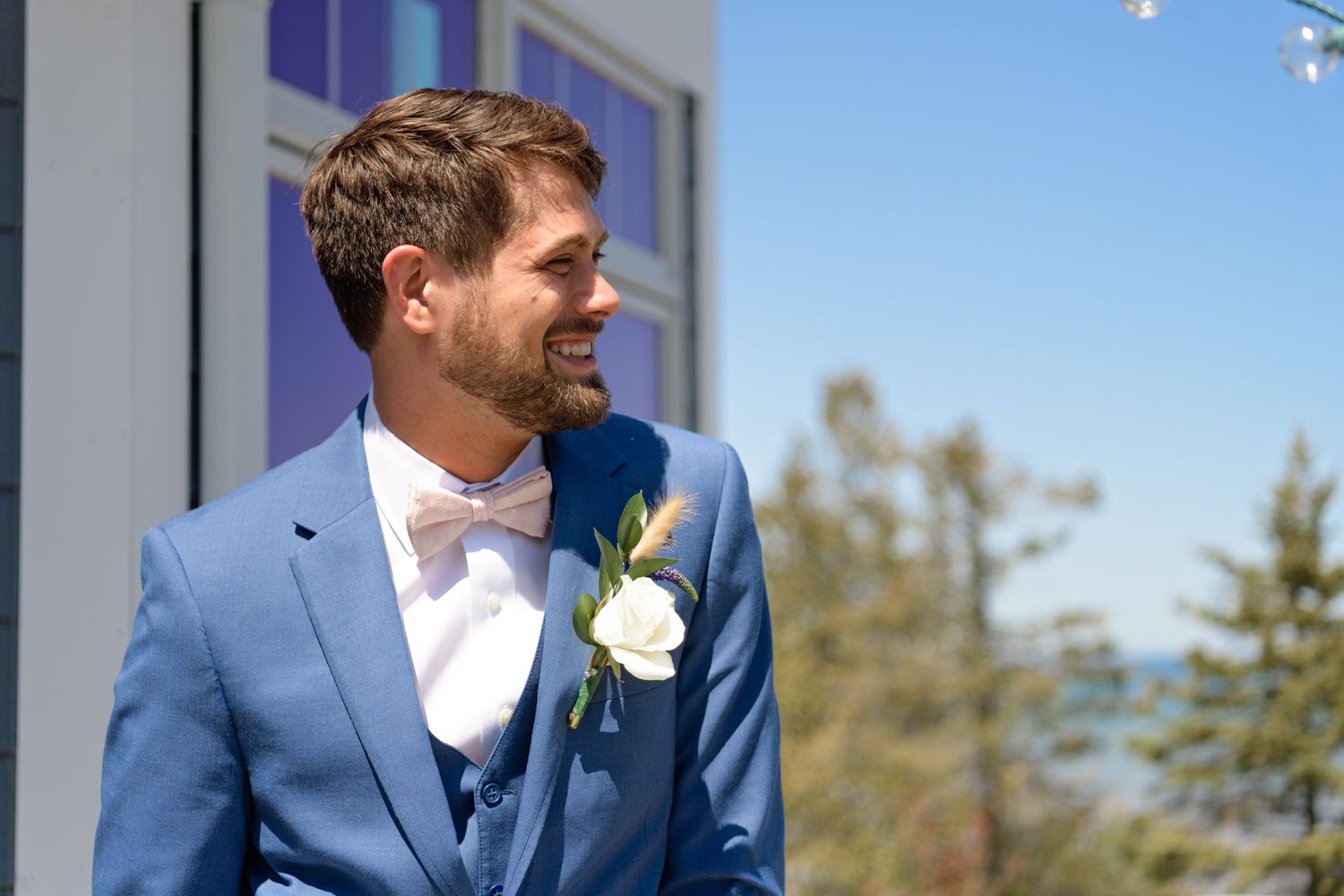 A groom in a blue suit and bow tie smiles outdoors on his wedding day.