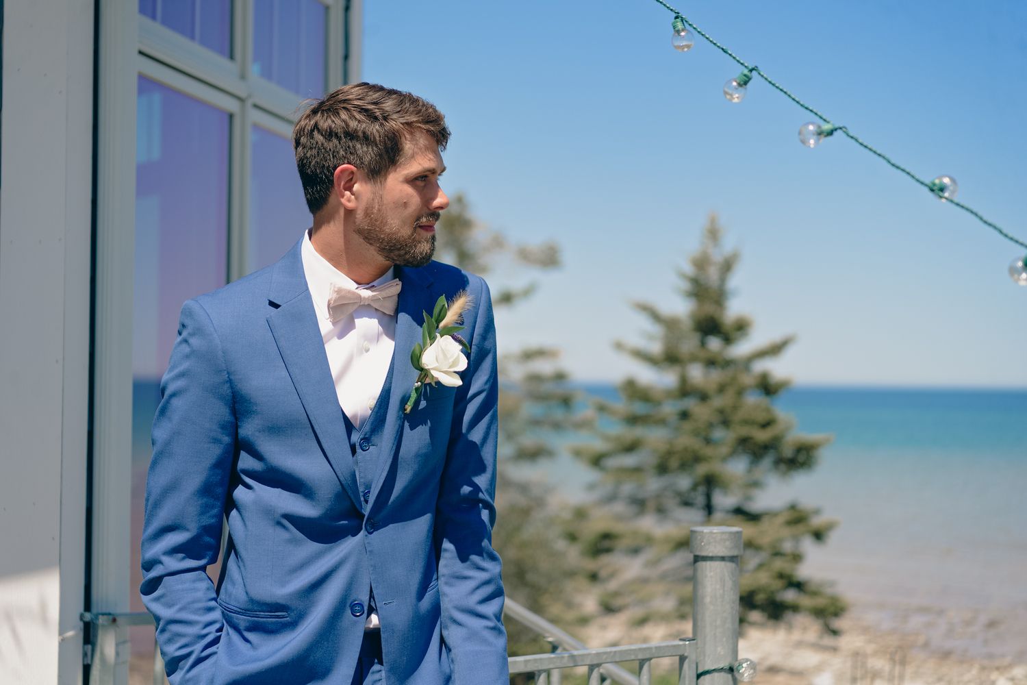 A groom in a blue suit with white boutonniere stands on a deck overlooking a lake.