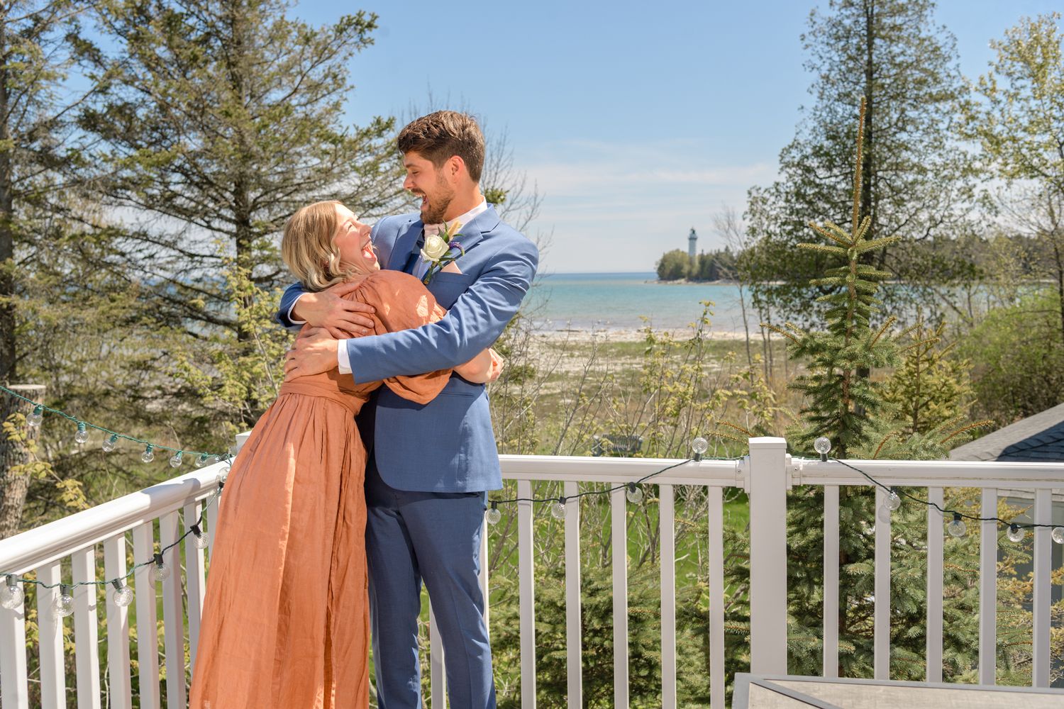 A couple embraces on a white deck overlooking a scenic lake and pine trees.