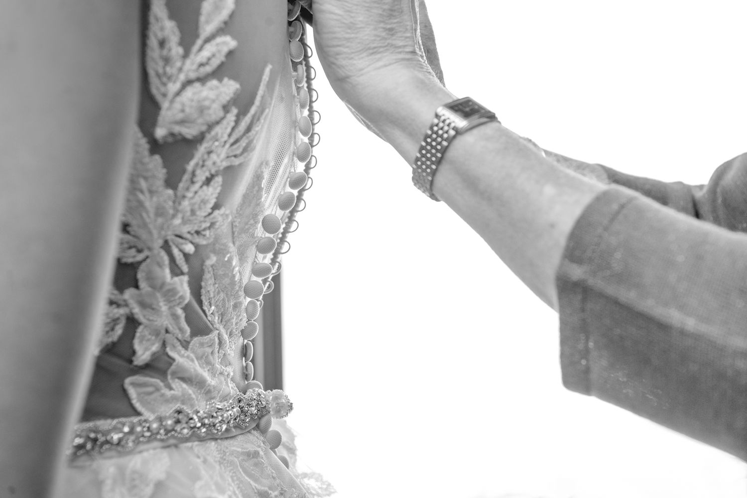 Black and white image of hands buttoning up a detailed wedding dress.