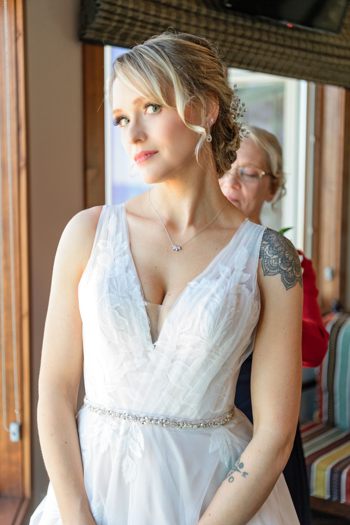 A bride wearing a white V-neck wedding gown with beaded belt stands by a window.