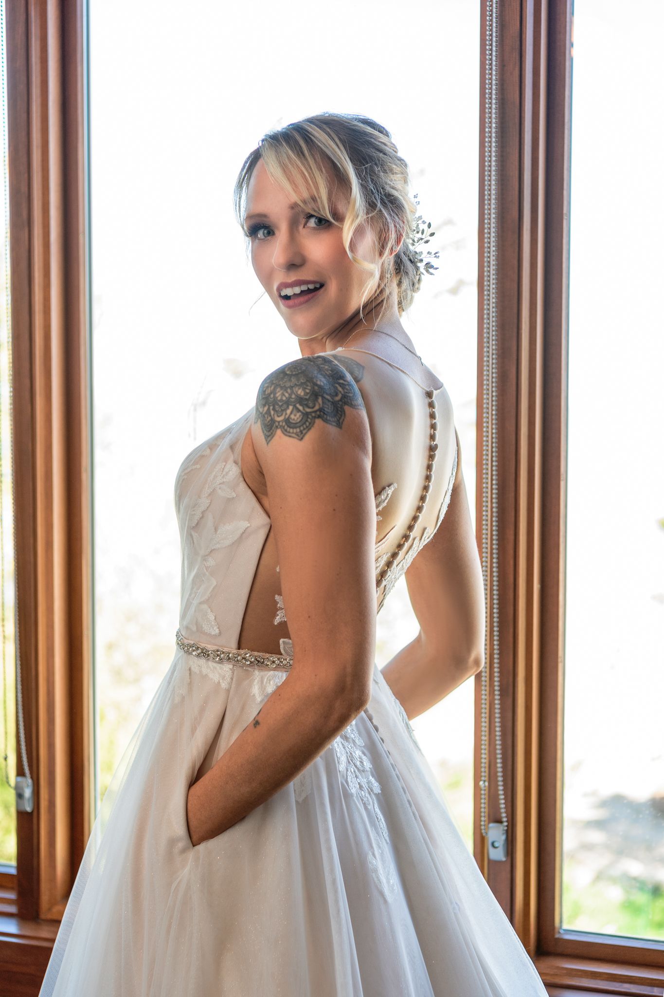 A bride in a white wedding gown with pockets poses near wooden-framed windows, showing off her shoulder tattoo.