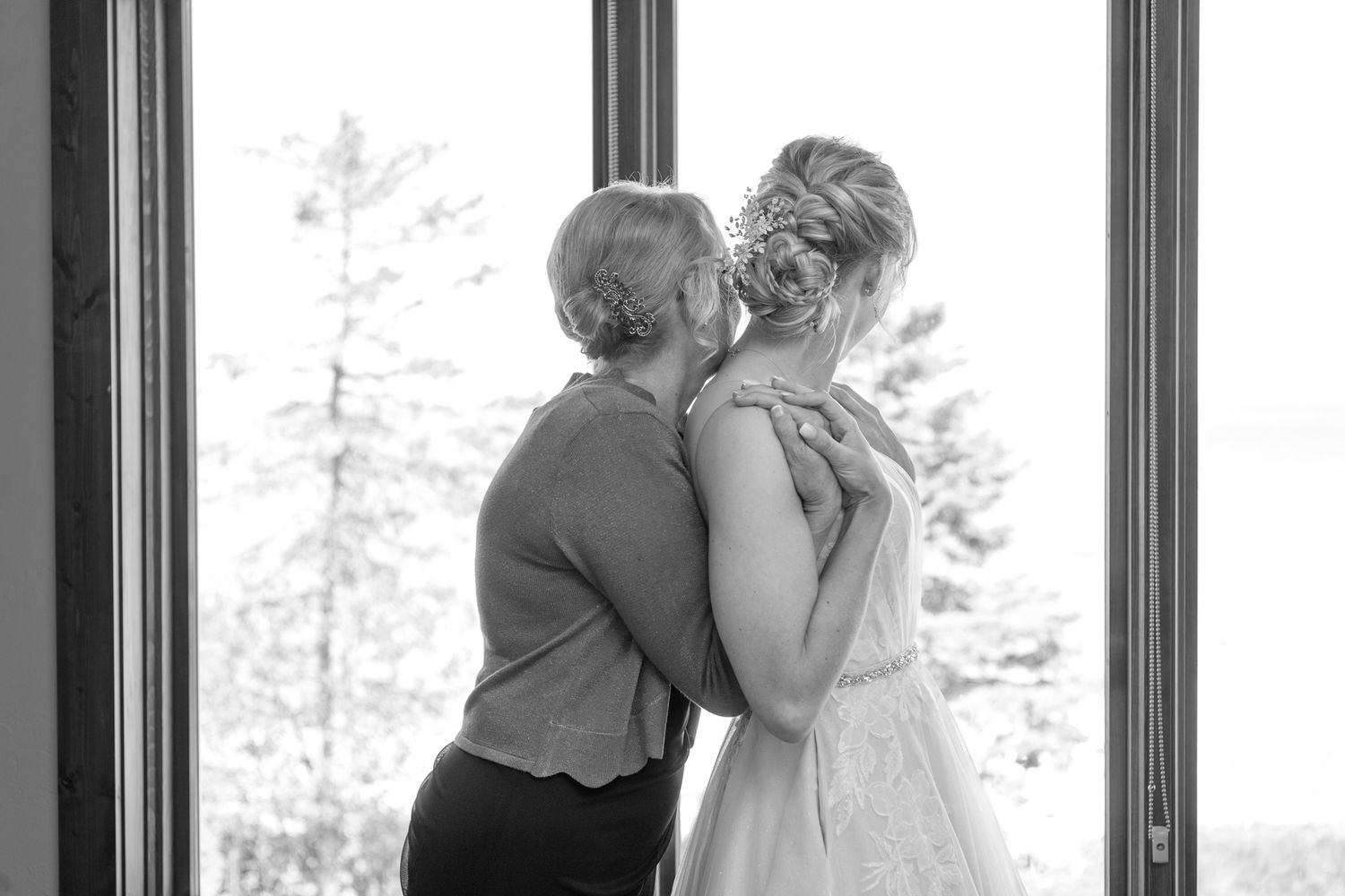 Black and white intimate moment captured by window with evergreen trees visible outside.